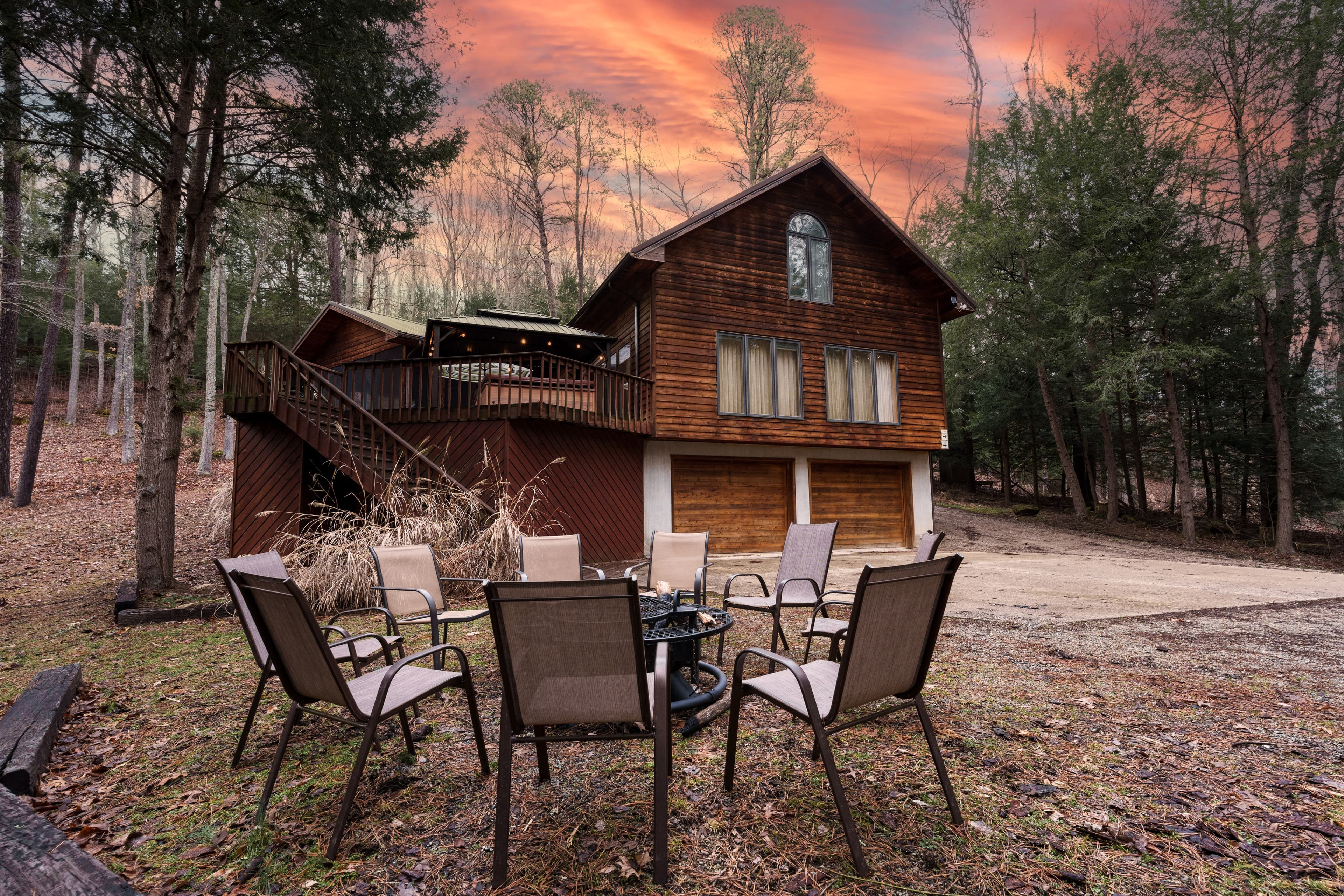 A wooden cabin surrounded by trees, featuring a seating area with chairs and a table in the foreground, under a colorful sunset sky.