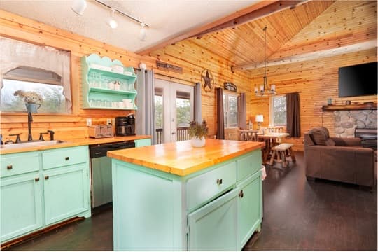 A cozy kitchen with a mint green island and wooden accents, featuring a living area with a stone fireplace.