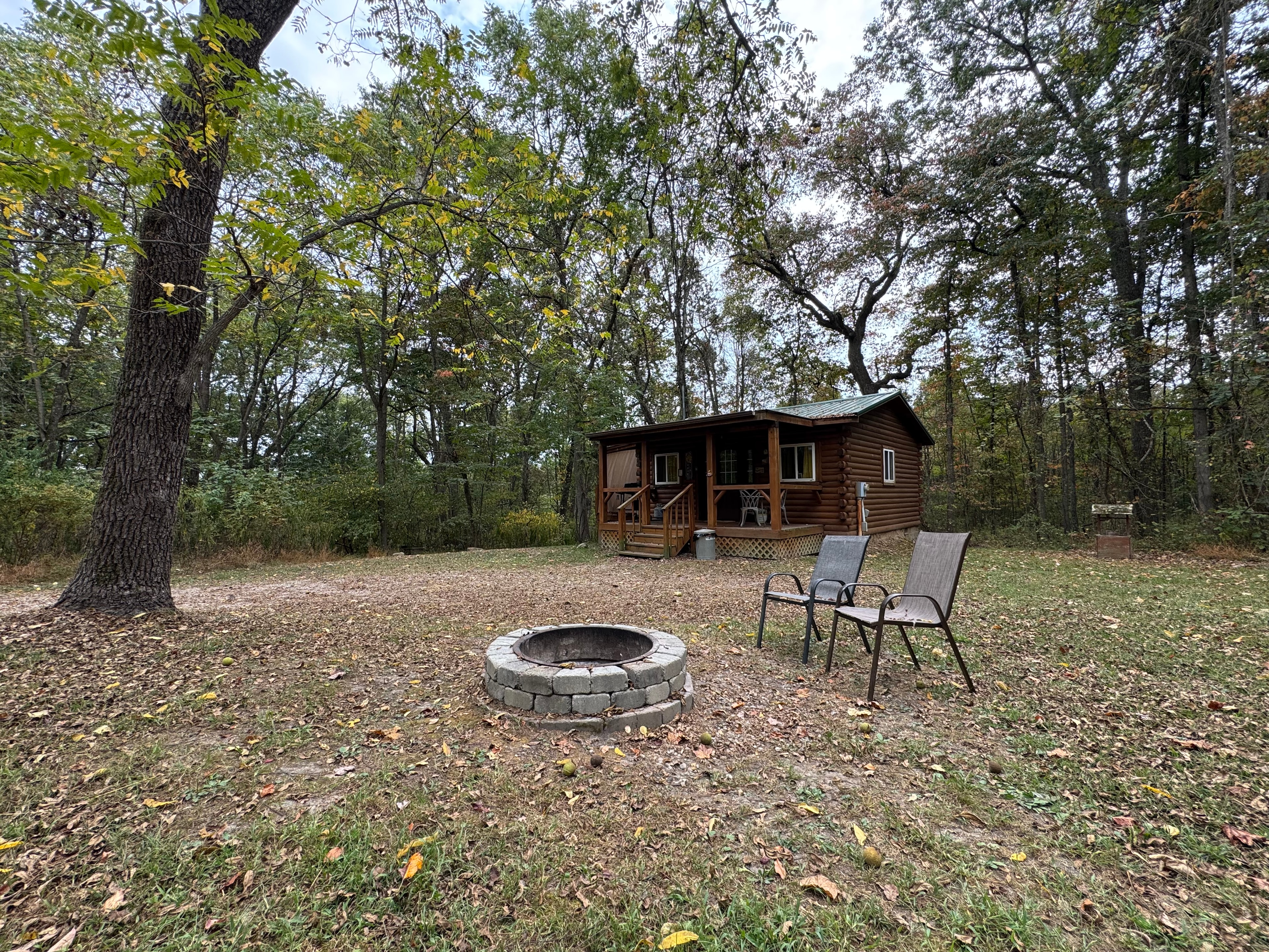 A rustic cabin surrounded by trees, featuring a stone fire pit and two chairs in the foreground.