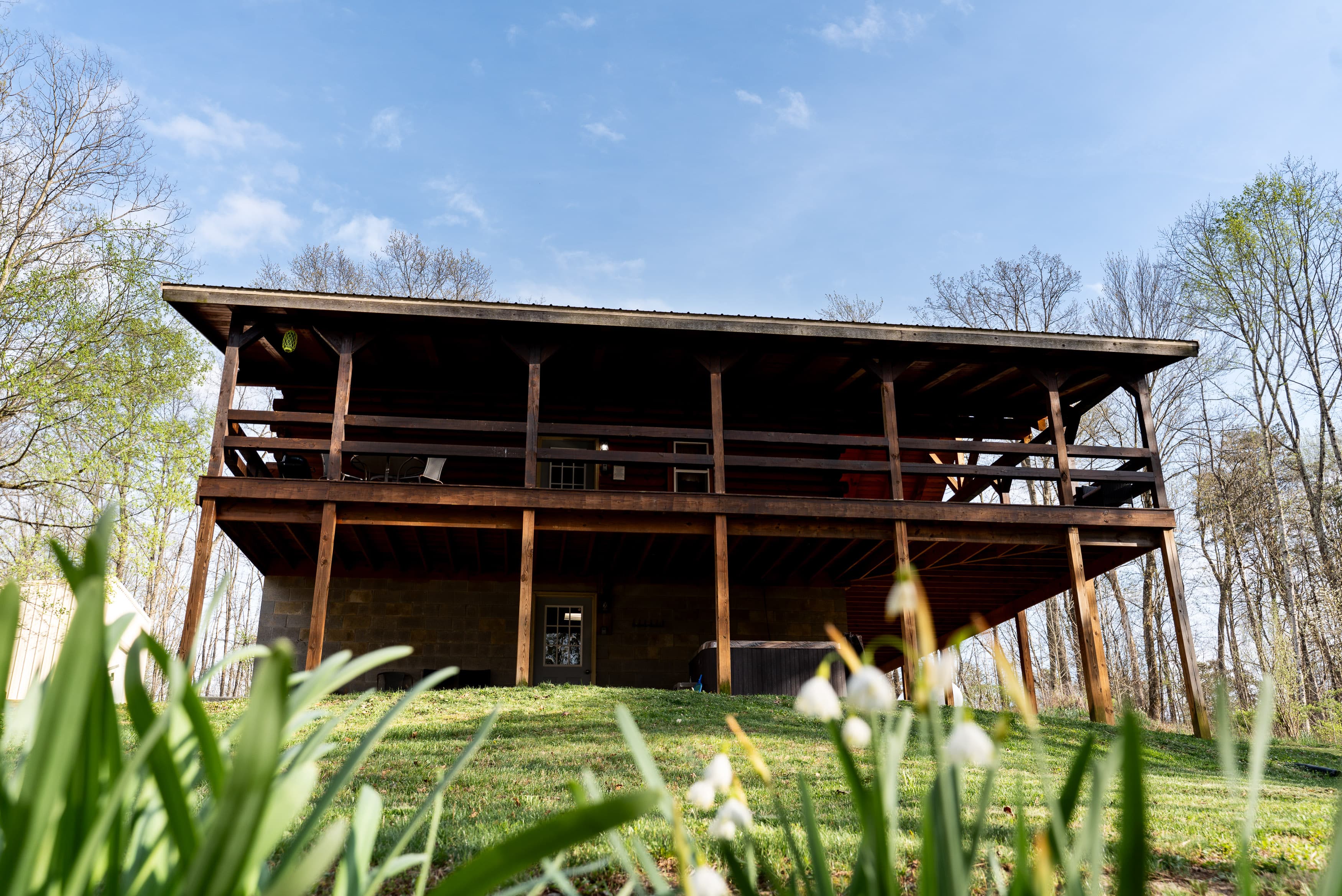 A wooden house on stilts with a covered porch, surrounded by green grass and budding flowers under a blue sky.