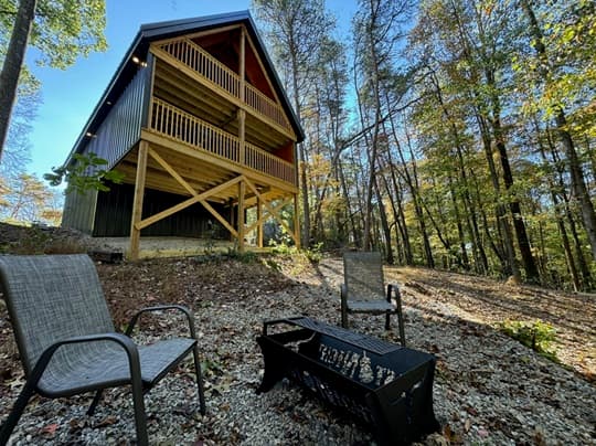 A modern cabin with a wooden deck surrounded by trees, featuring two chairs and a fire pit in the foreground.