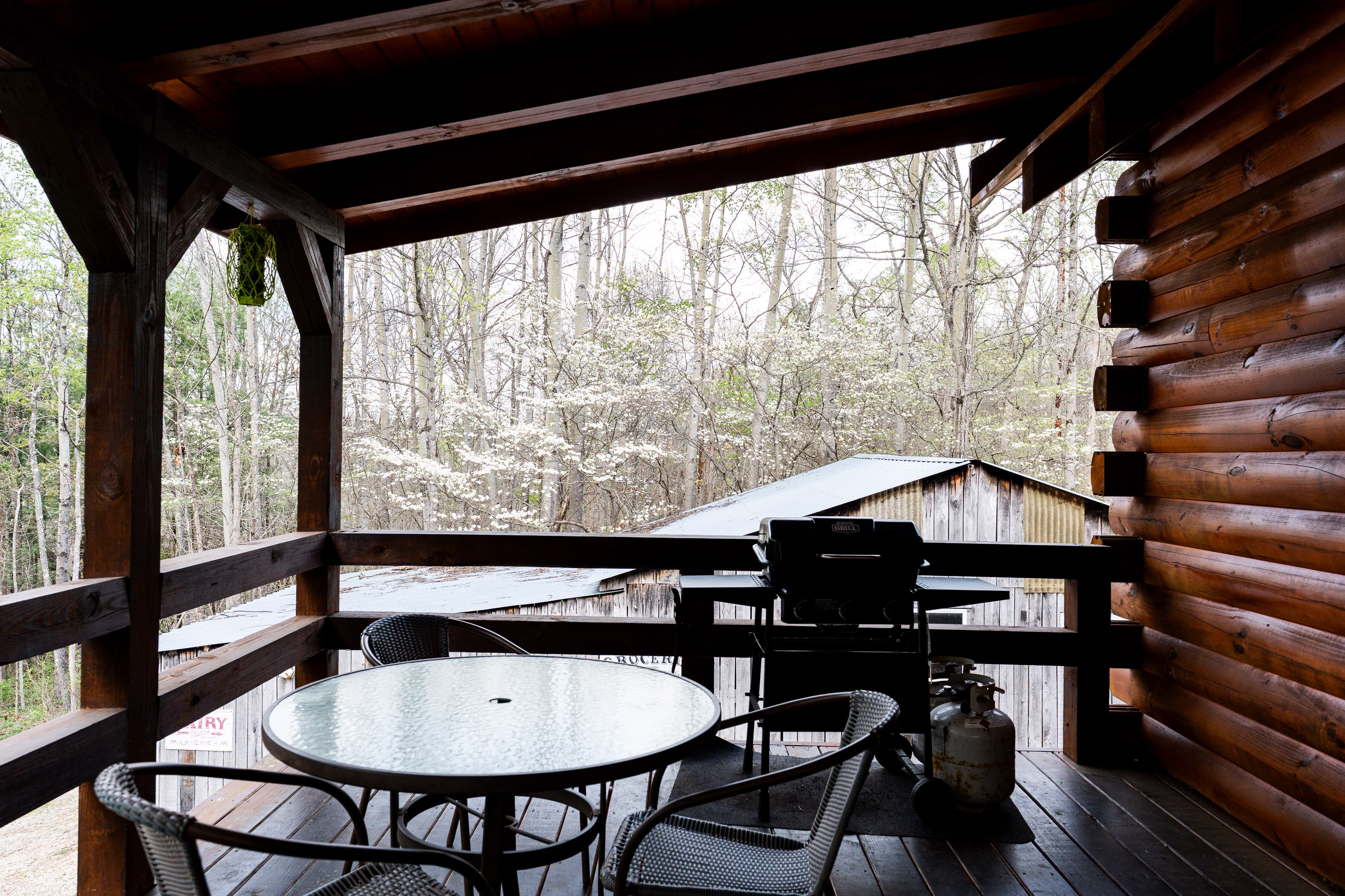 A wooden porch with a glass table, chairs, and a grill overlooks a wooded area.