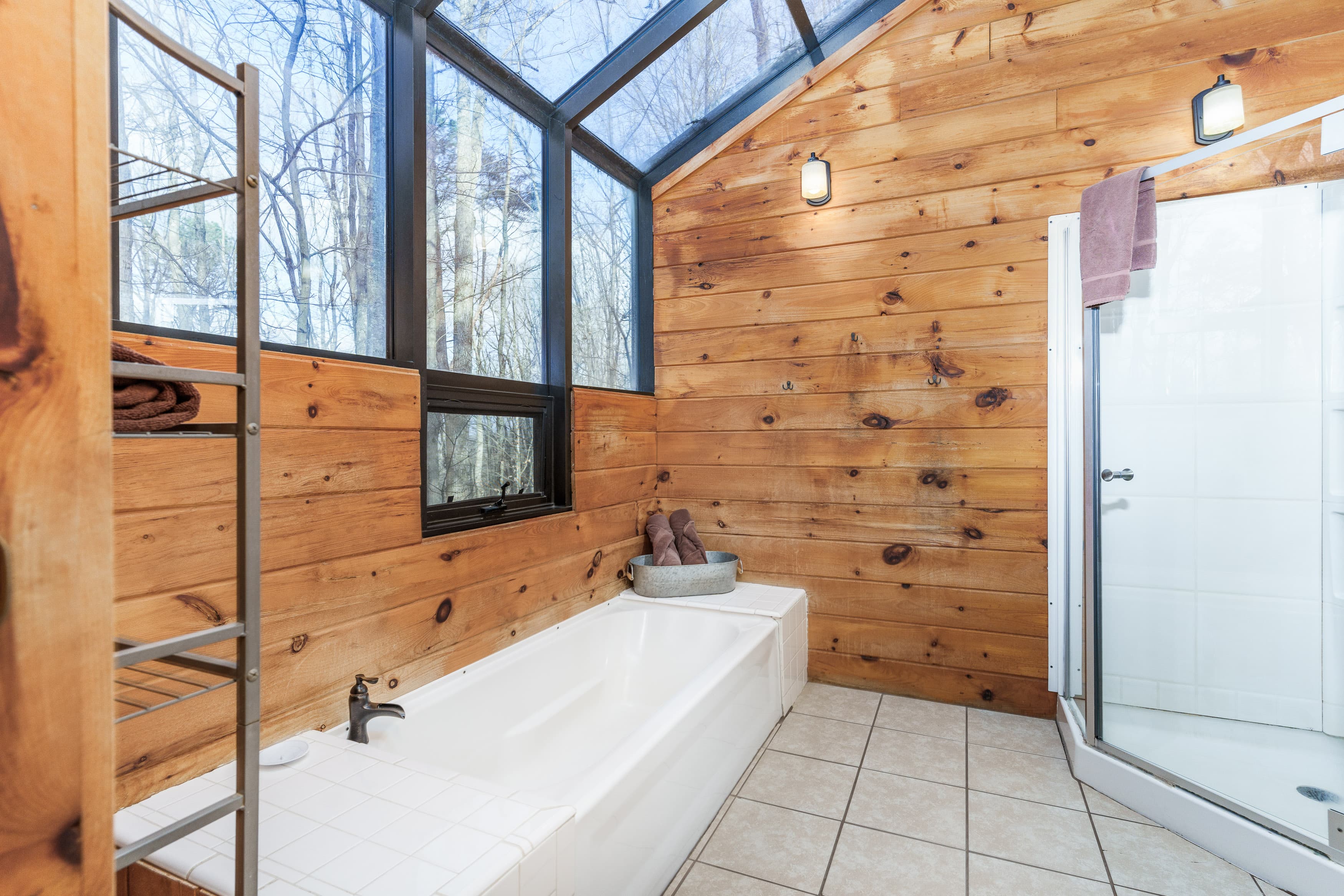 Bright, wooden-paneled bathroom featuring a bathtub, shower, and large windows with a view of trees.