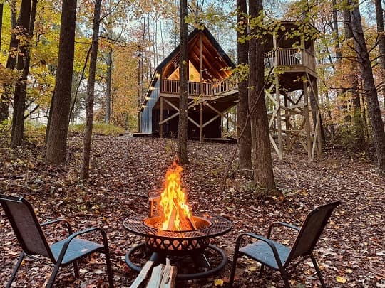 A cozy campfire sits in front of a treehouse surrounded by autumn foliage.