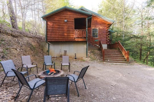 A rustic log cabin surrounded by trees, featuring a fire pit and seating area in front.