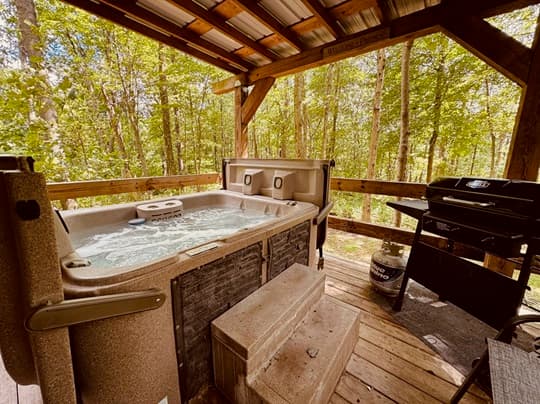 Outdoor hot tub surrounded by trees with a grill nearby.