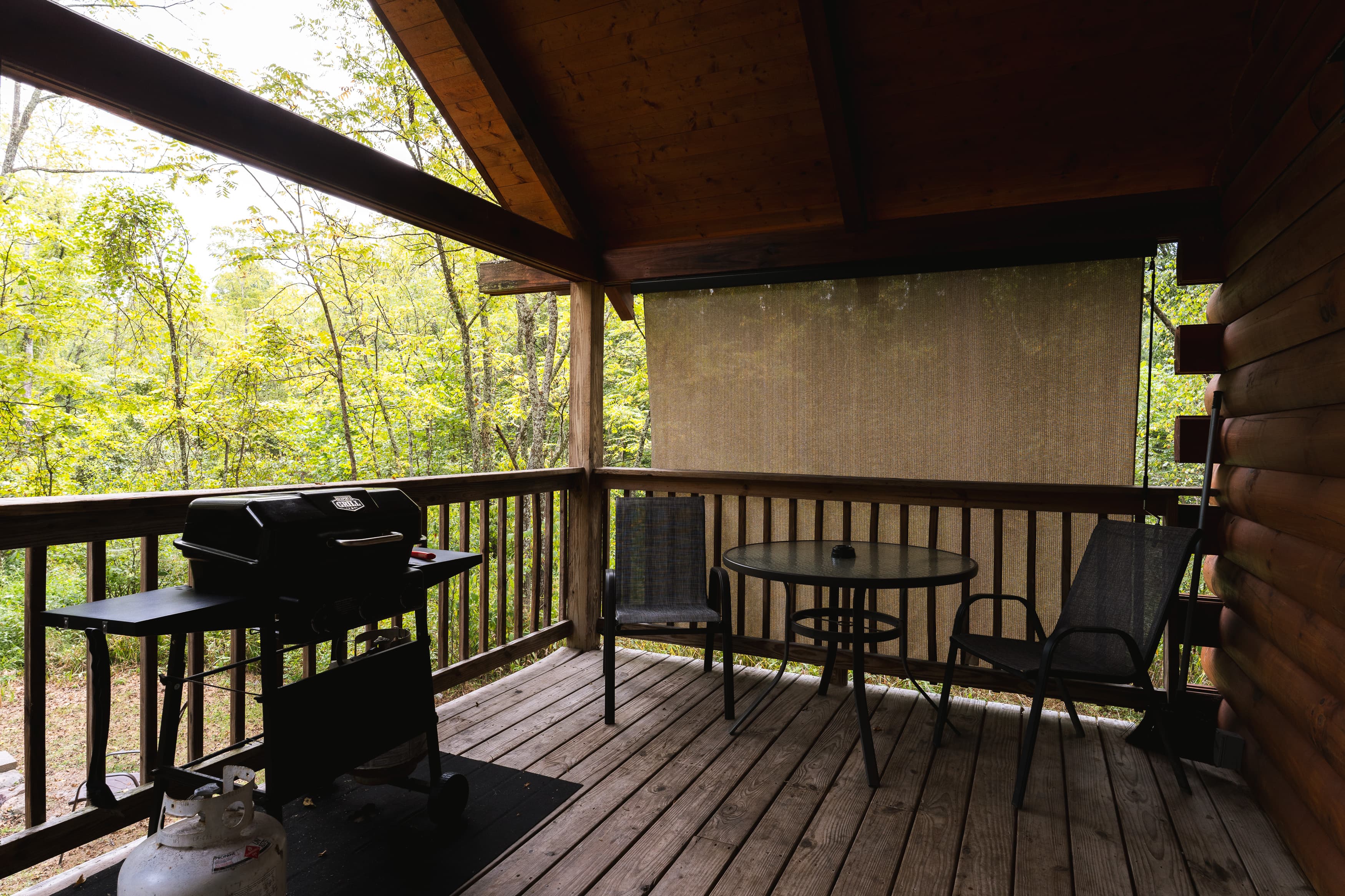 A covered outdoor deck with a grill, a table, and chairs overlooks a wooded area.