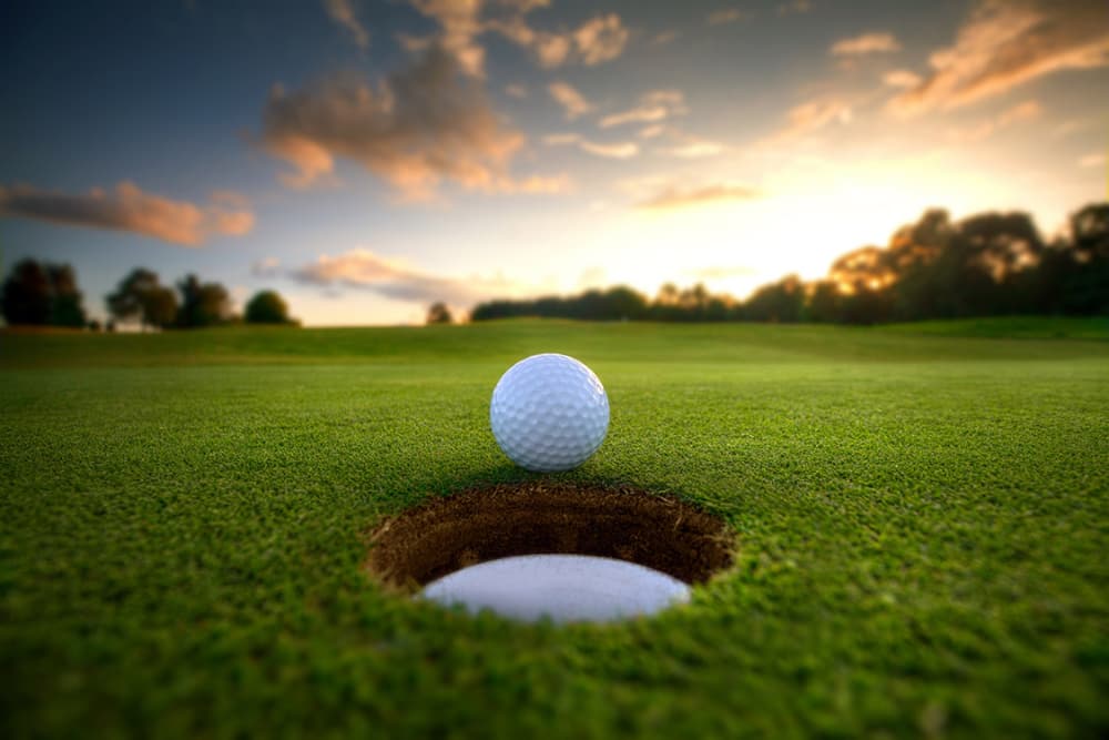 A golf ball is poised near a hole on a lush green golf course at sunset.