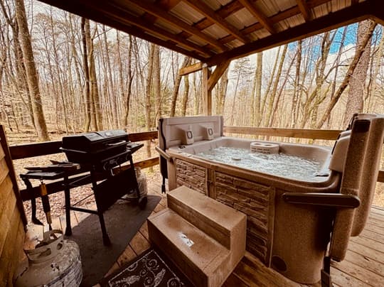 A hot tub with bubbling water is situated on a wooden deck beside a grill, surrounded by a serene forest.