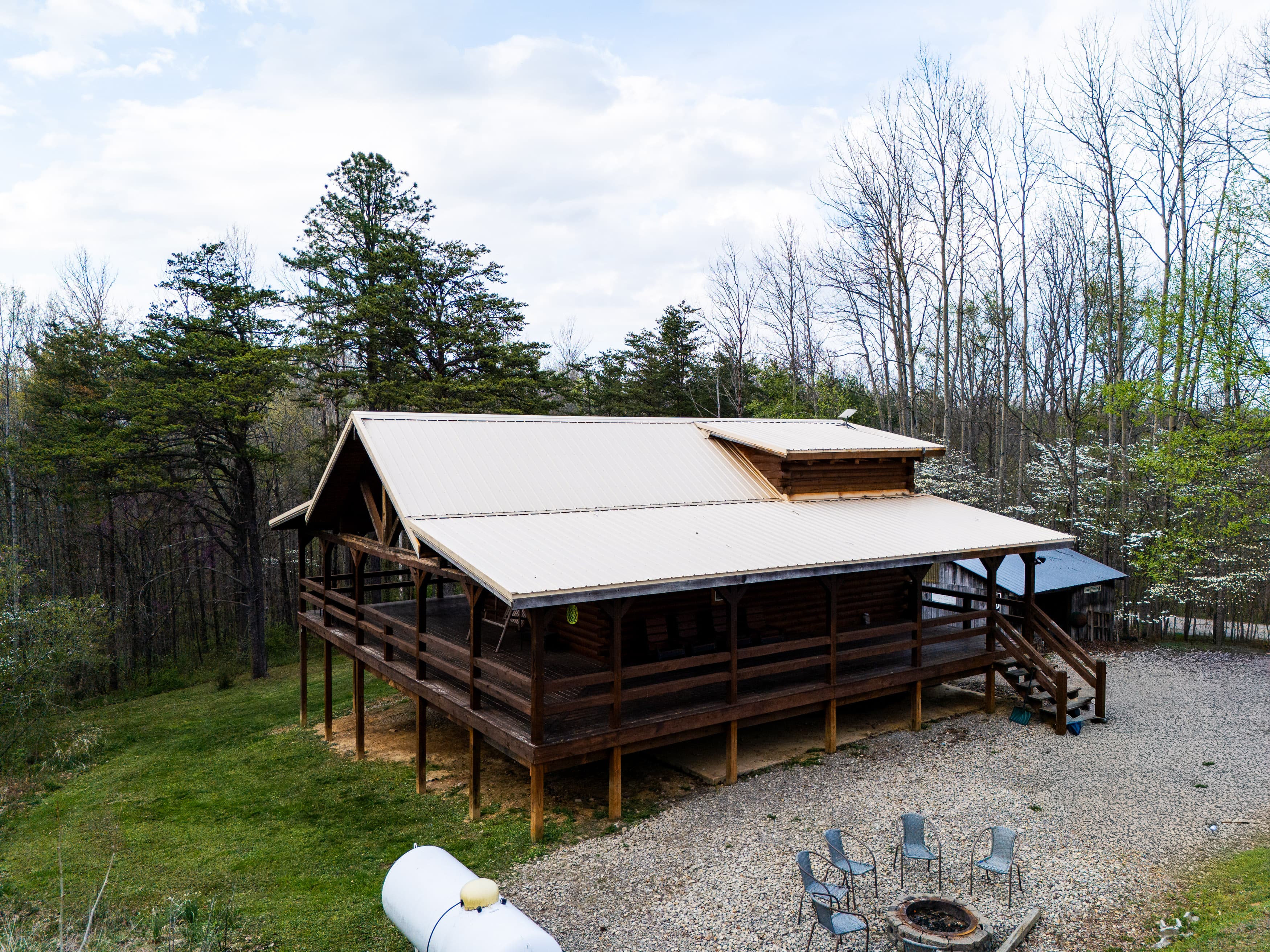 Aerial view of a log cabin with a wraparound porch surrounded by trees and a gravel fire pit area.