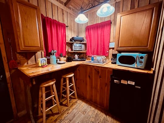 Cozy wooden kitchen with red curtains, a sink, a small refrigerator, and two bar stools.