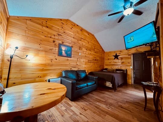 Cozy wooden interior of a cabin, featuring a leather couch, bed, table, and a wall-mounted TV.