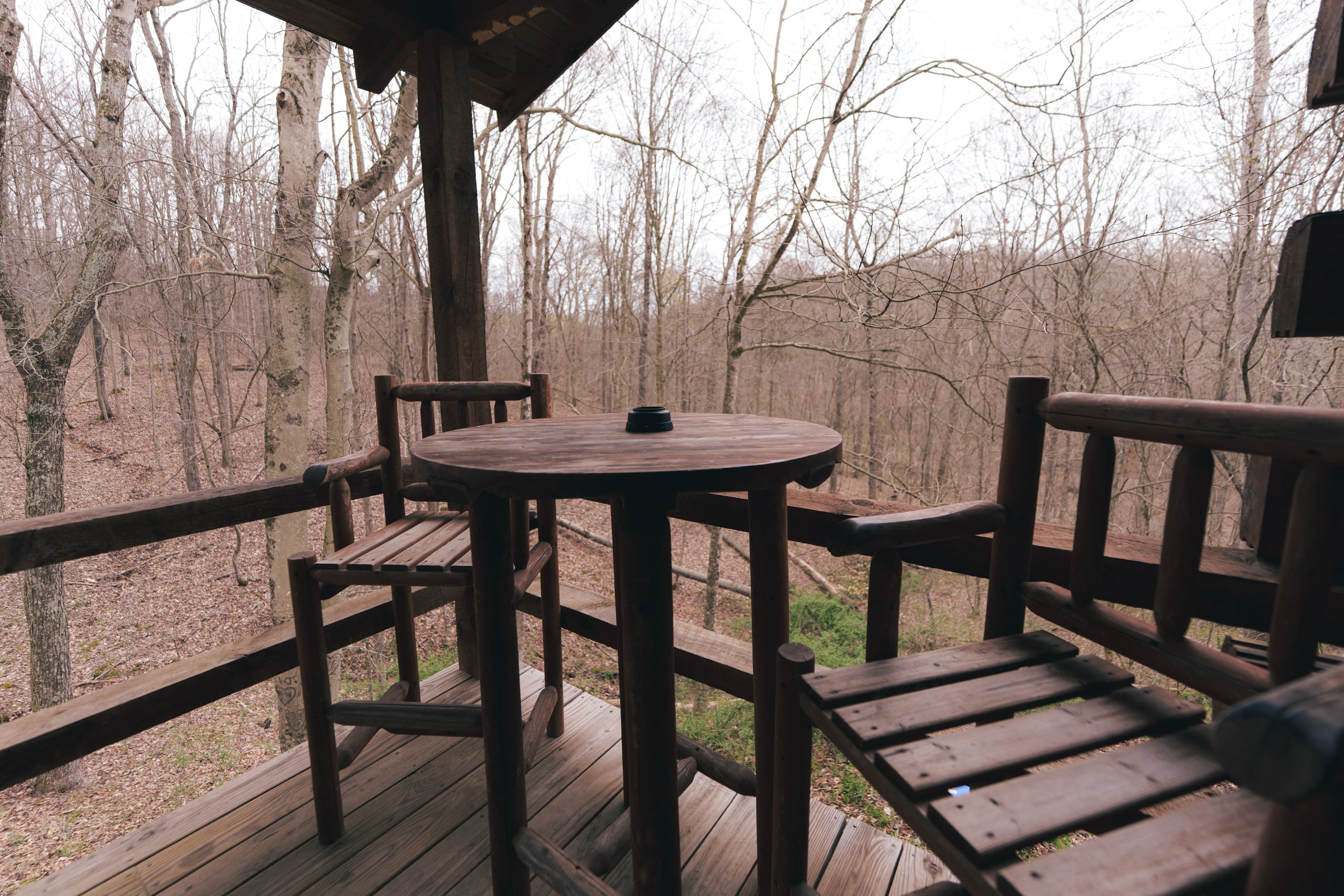 A wooden balcony with two chairs and a round table overlooks a bare, wooded landscape.