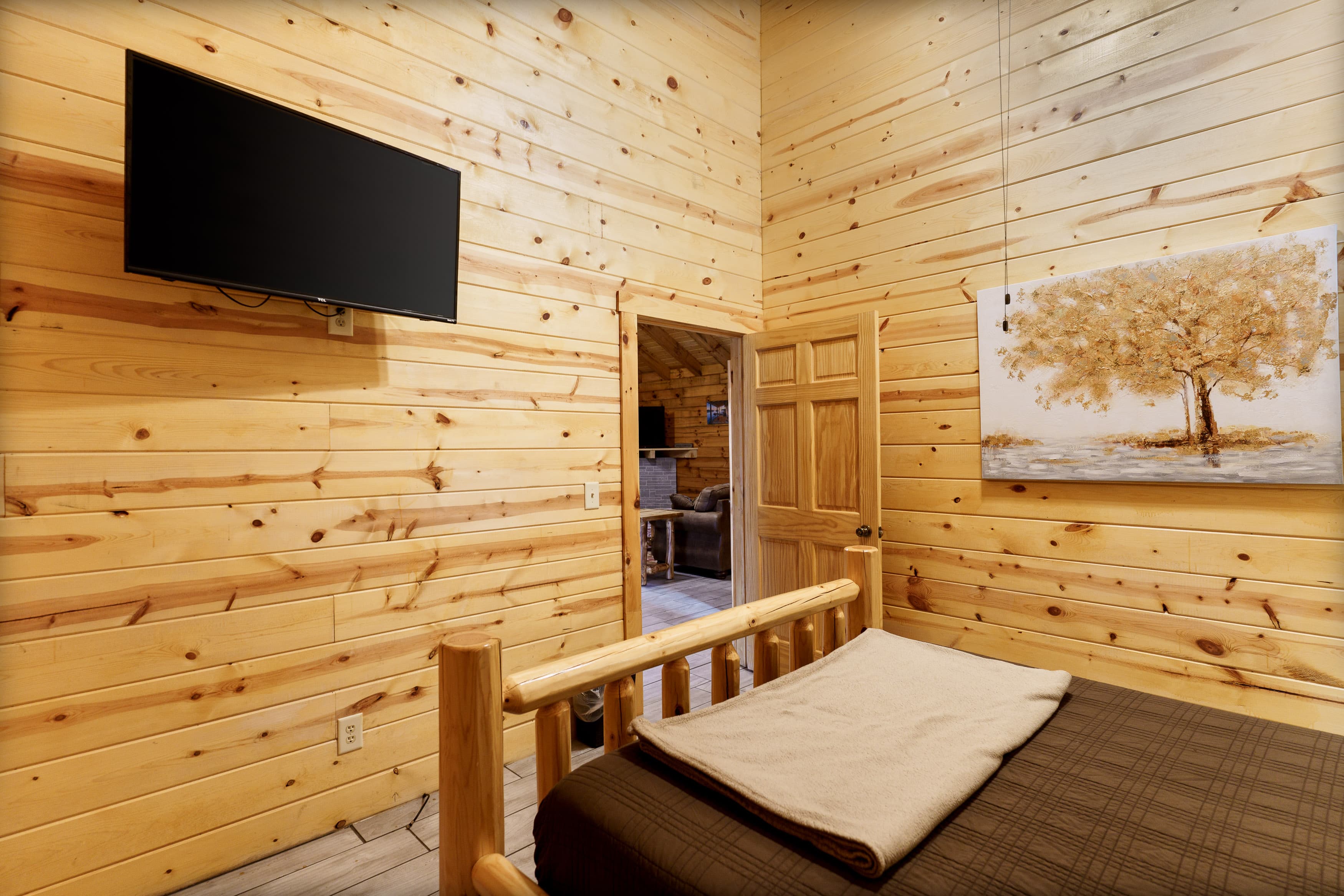 Cozy wooden bedroom featuring a TV, a bed with a blanket, and a wall art of a tree.