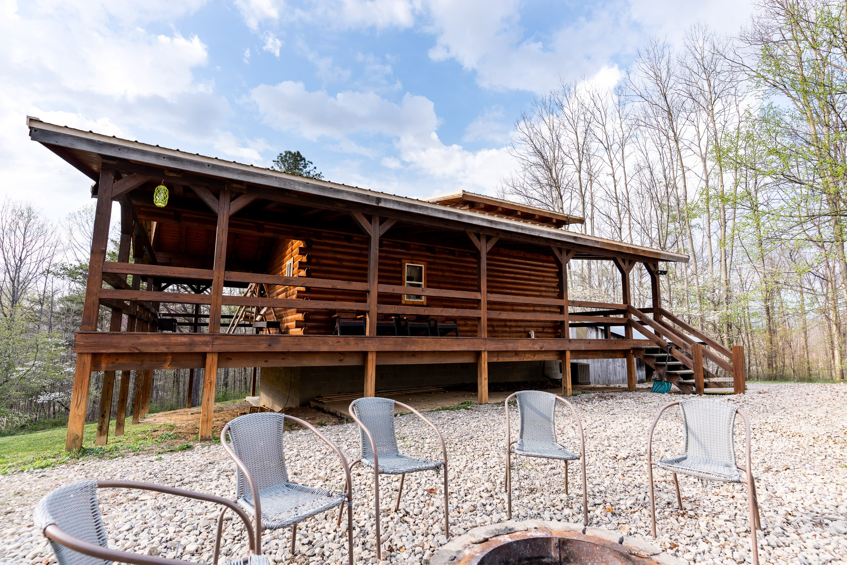 A rustic log cabin with a front porch overlooks a fire pit surrounded by chairs, set amidst a wooded area.