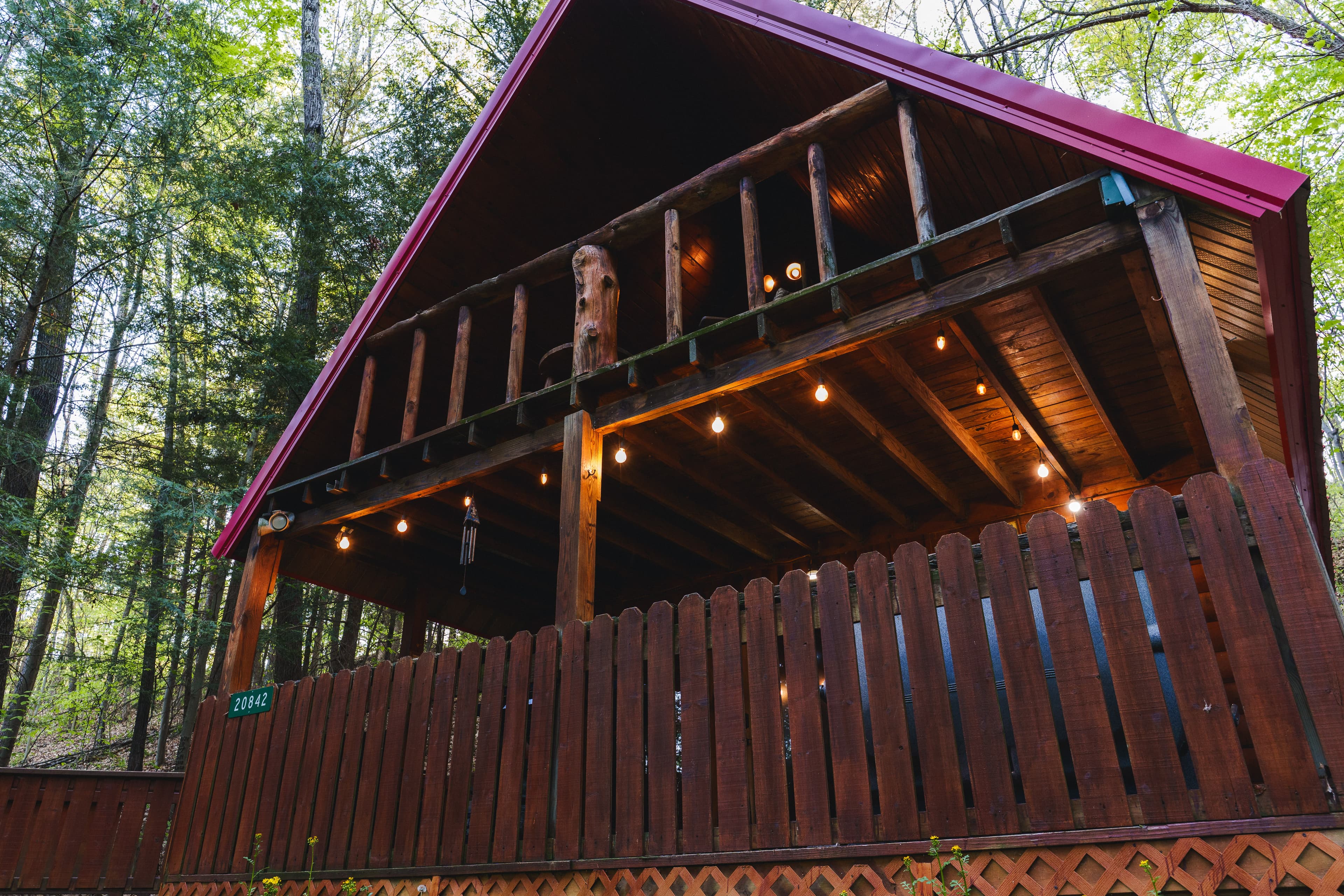 A rustic cabin with a pink roof, wooden railing, and string lights nestled among trees.