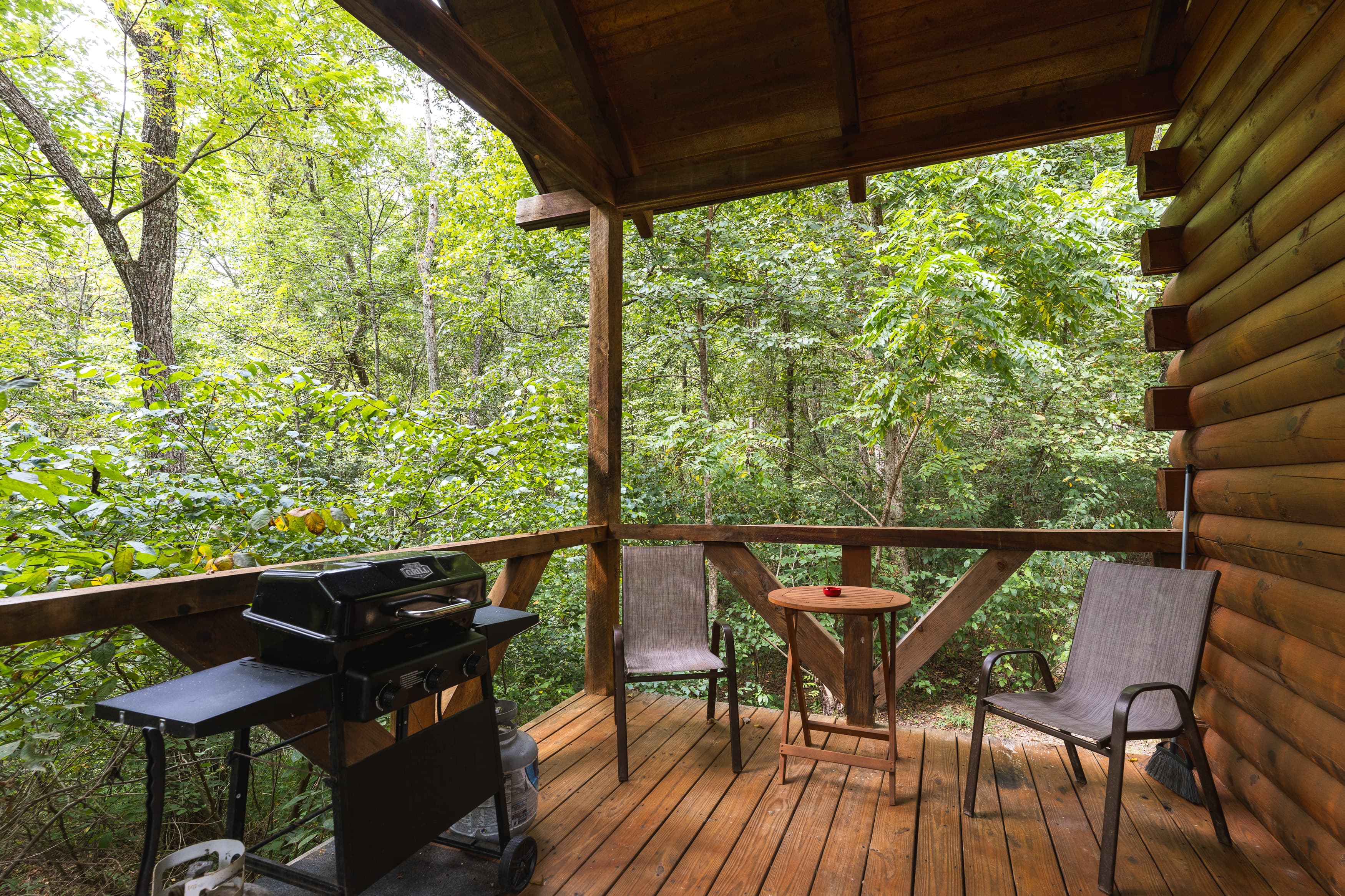 A cozy outdoor porch with a grill, two chairs, and a small table surrounded by lush greenery.