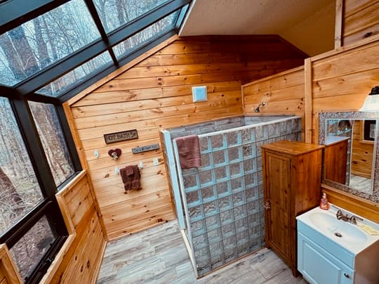 A wooden bathroom with a glass-block shower, a sink, and large windows overlooking trees.