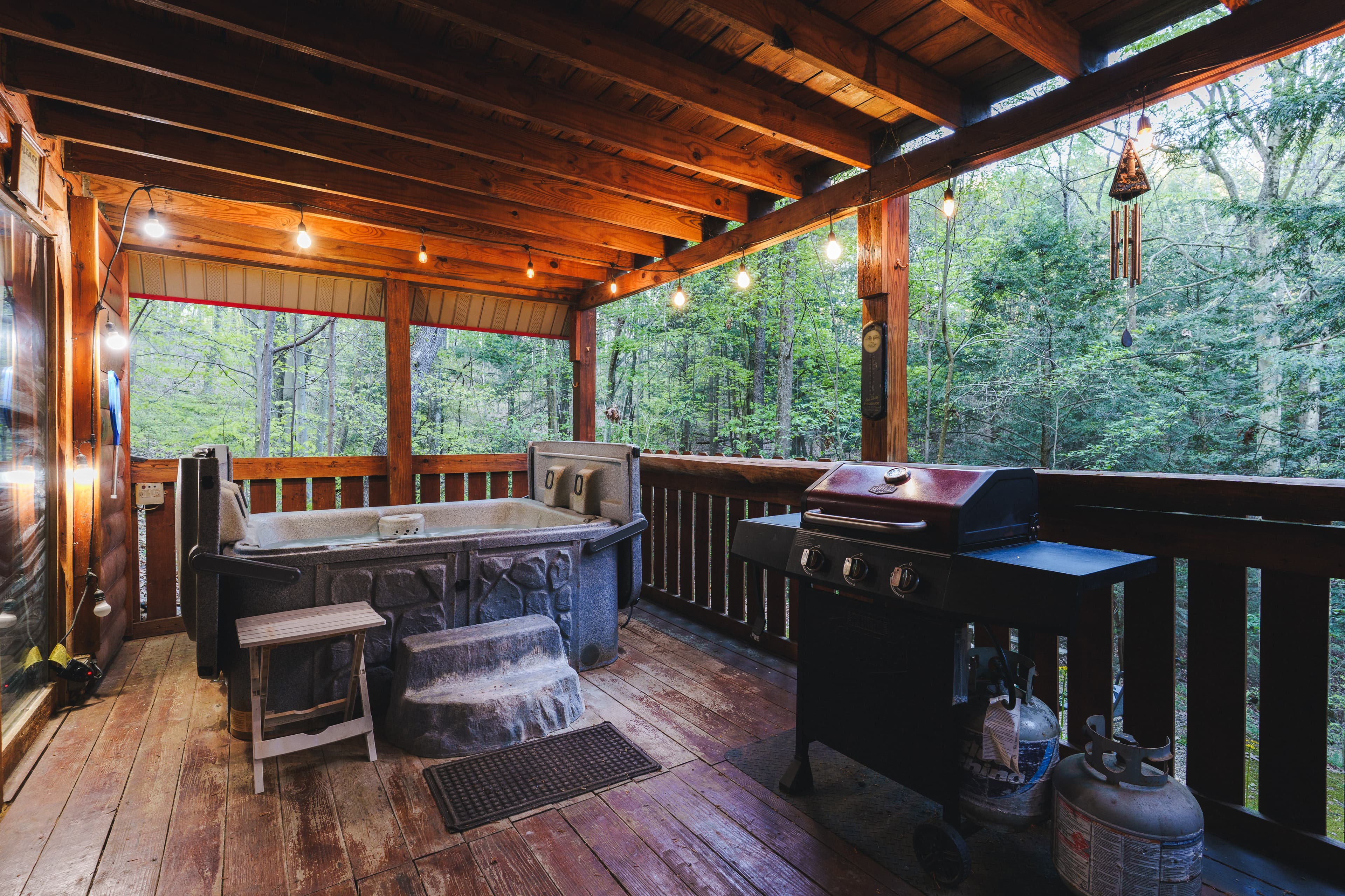 A cozy wooden deck featuring a hot tub, grill, and string lights, surrounded by lush greenery.