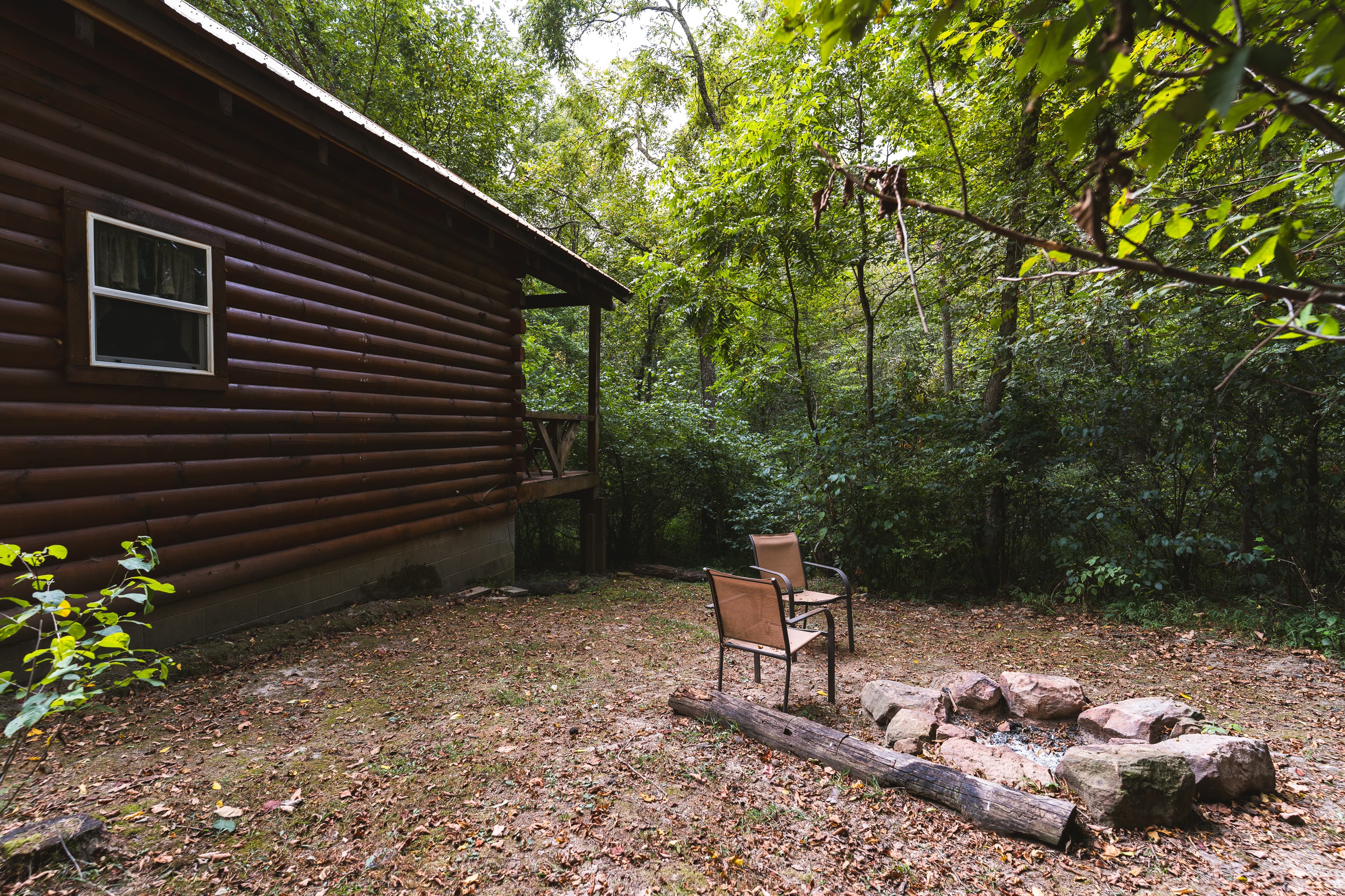 A rustic cabin in a wooded area with two chairs and a stone fire pit.