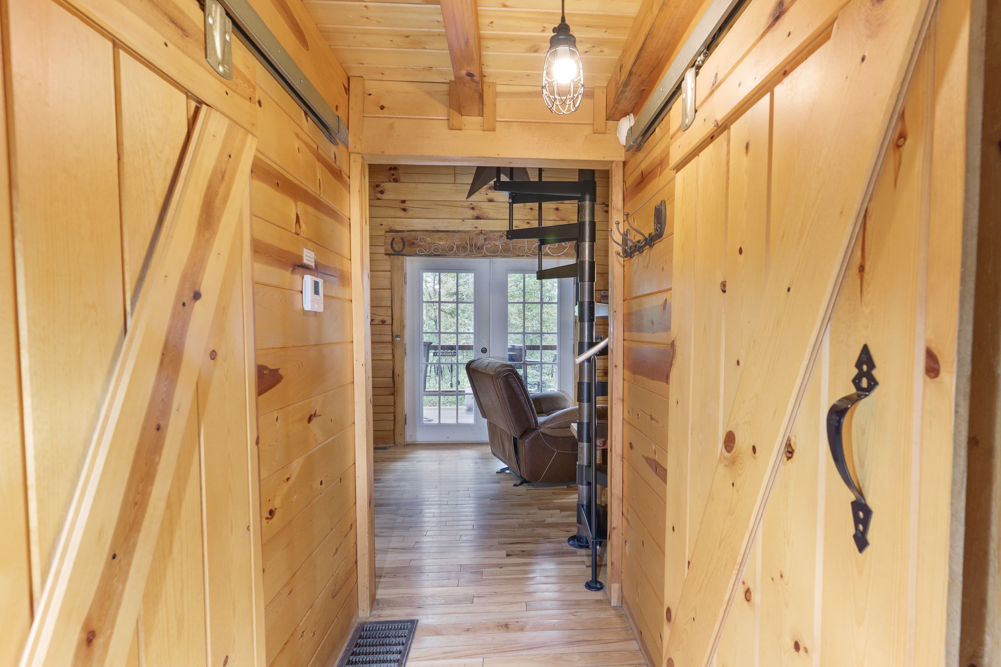 A rustic hallway with wooden walls and sliding barn doors leads to a cozy living area with a spiral staircase.