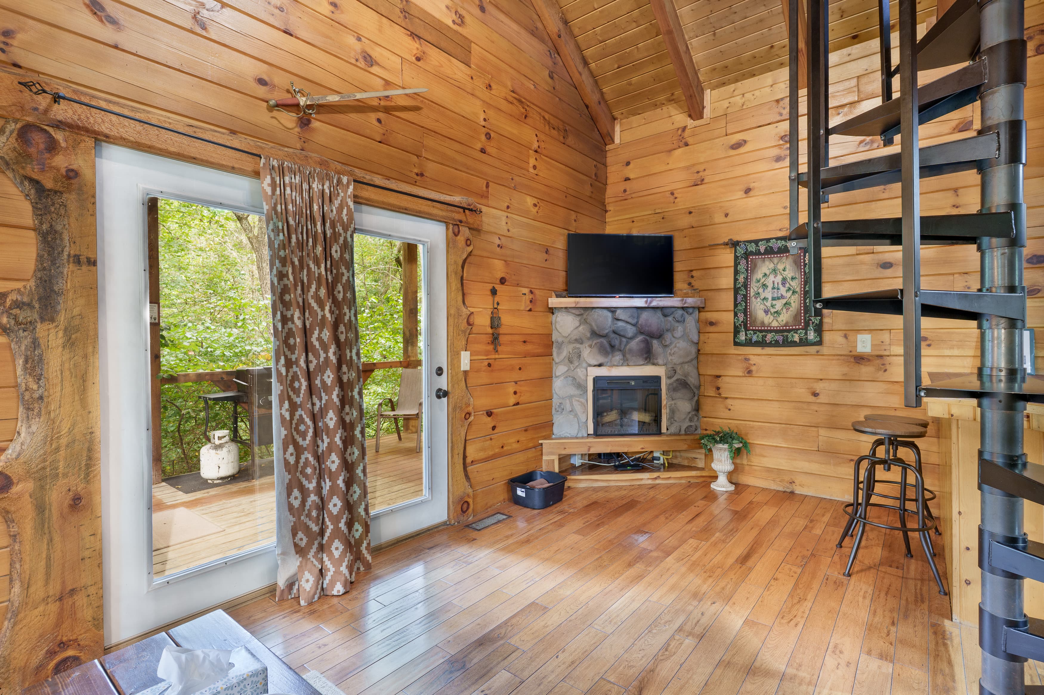 Cozy wooden interior with a spiral staircase, fireplace, and large windows offering a view of greenery.