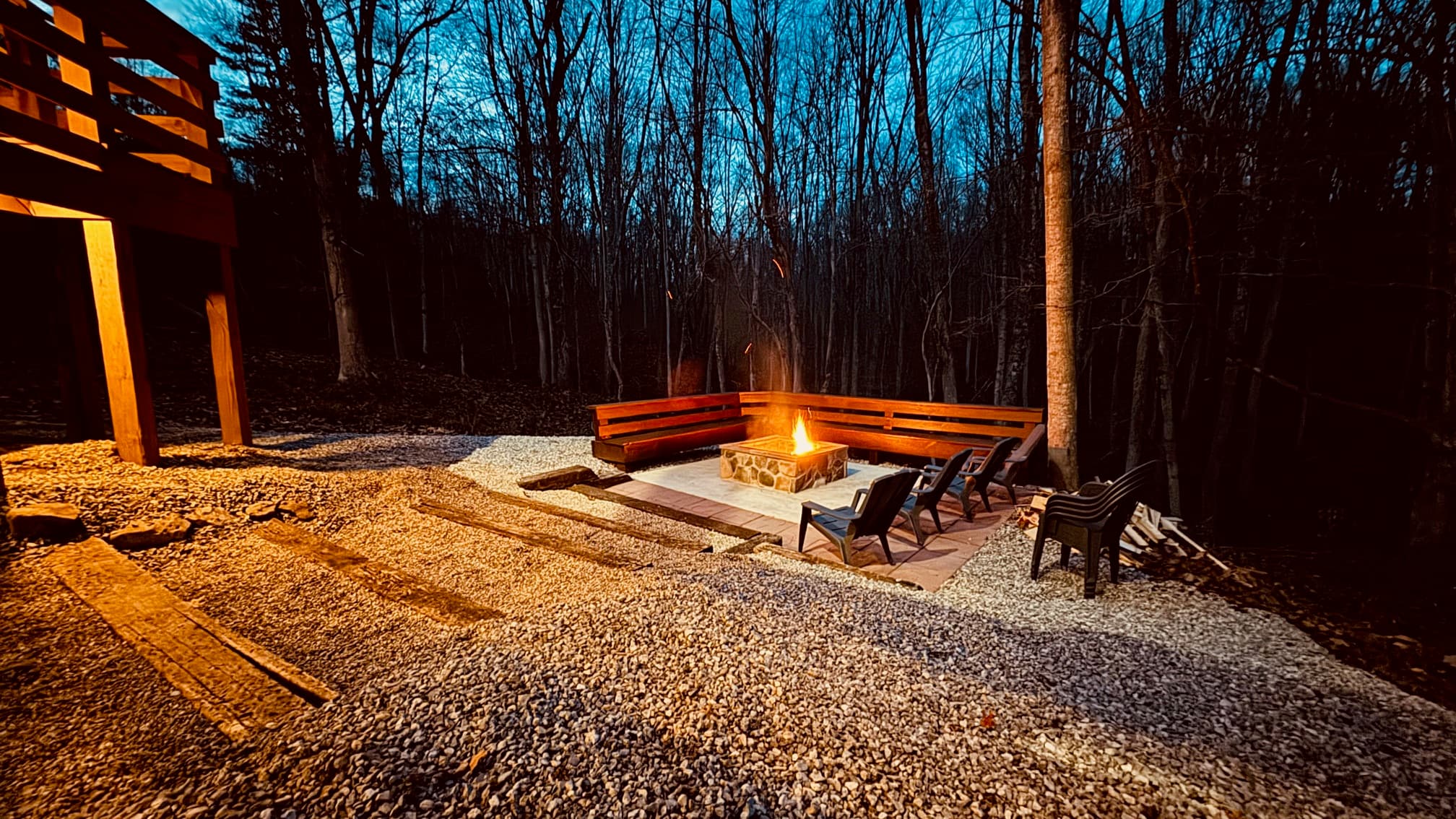 A fire pit surrounded by chairs in a wooded area at dusk.