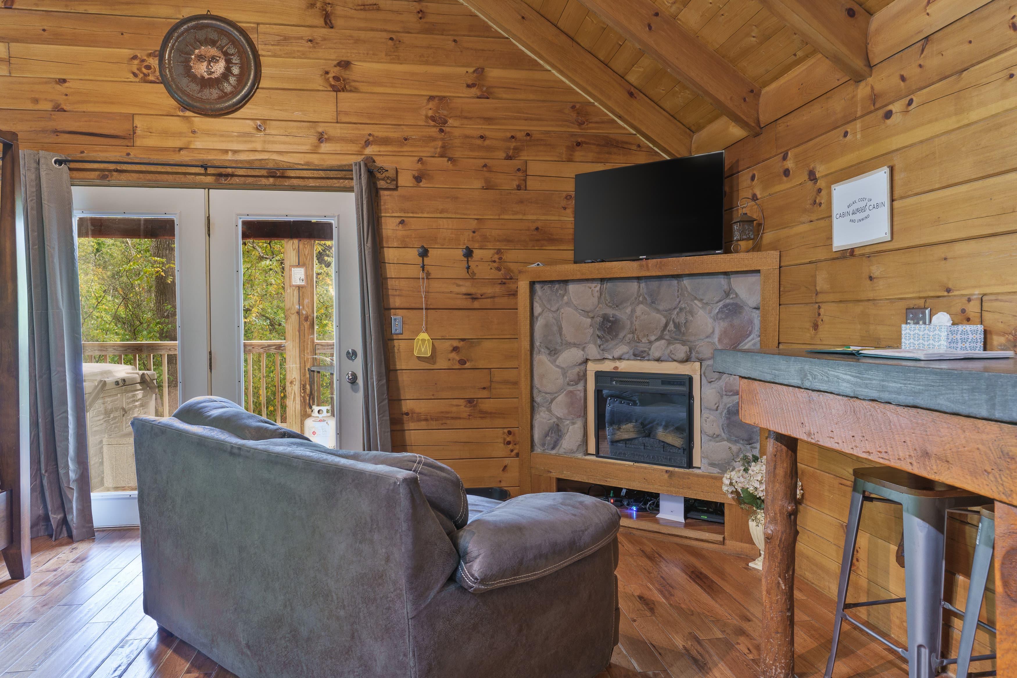 Cozy wooden cabin living room featuring a grey sofa, stone fireplace, and a television.