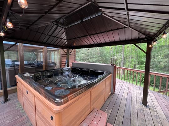 A wooden hot tub is situated under a gazebo on a deck surrounded by trees.