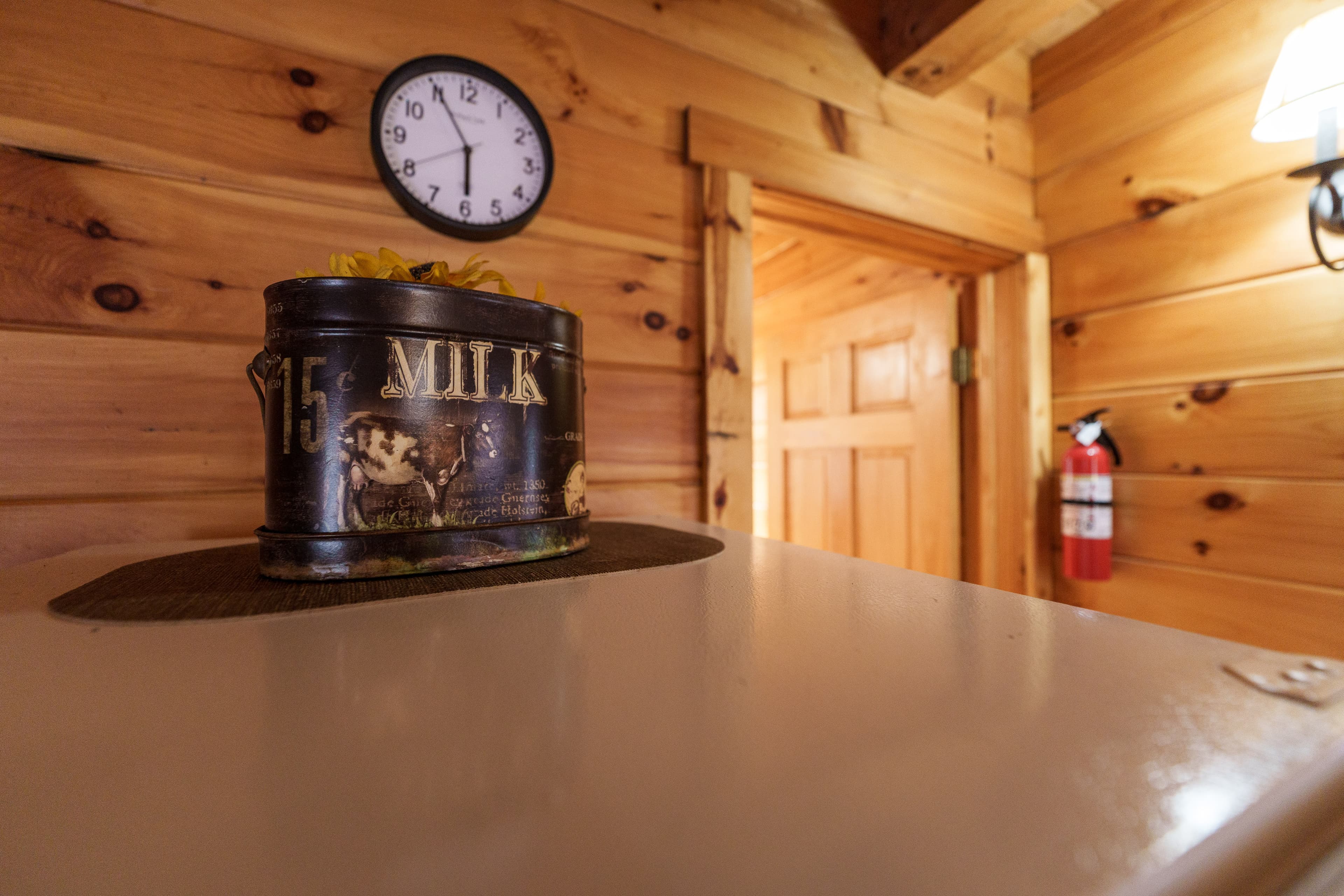 A vintage milk can with decorative flowers sits on a countertop in a wooden room, featuring a clock and a fire extinguisher in the background.