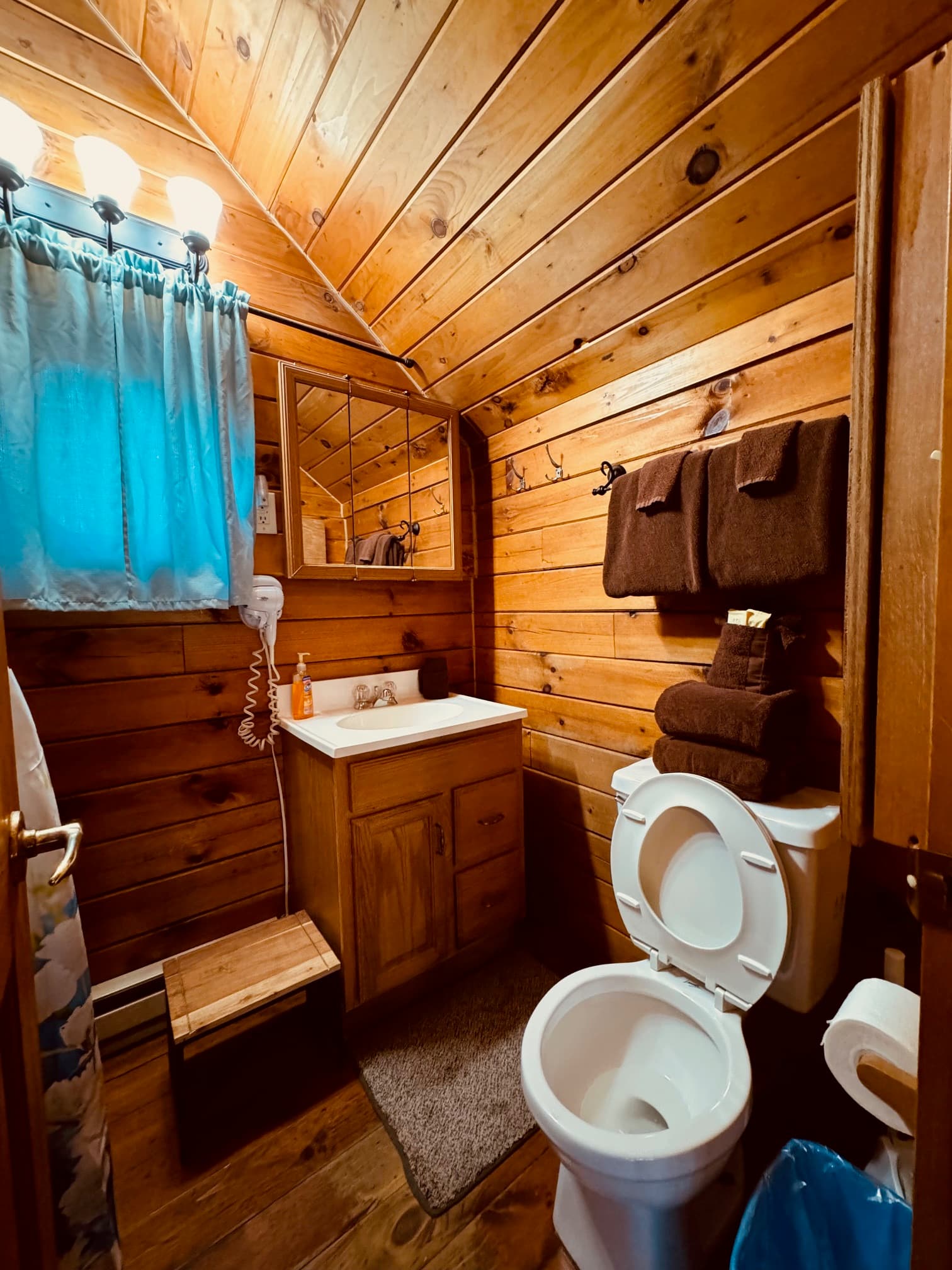 A cozy bathroom featuring wooden walls, a sink with a mirror, towels, and a toilet.