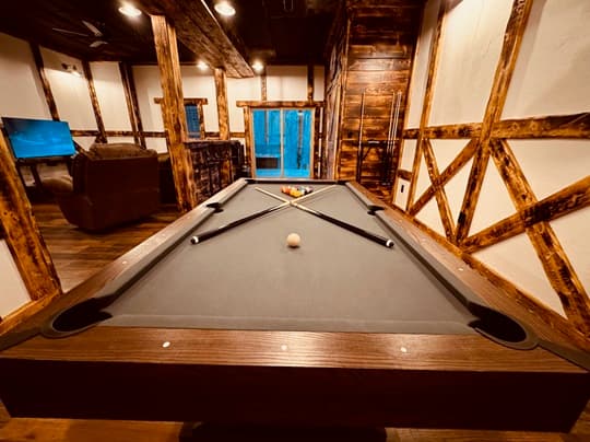 A pool table in a rustic room with wooden beams and a nearby window.