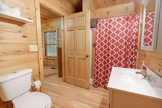A wooden-themed bathroom featuring a white toilet, sink, and a red patterned shower curtain.