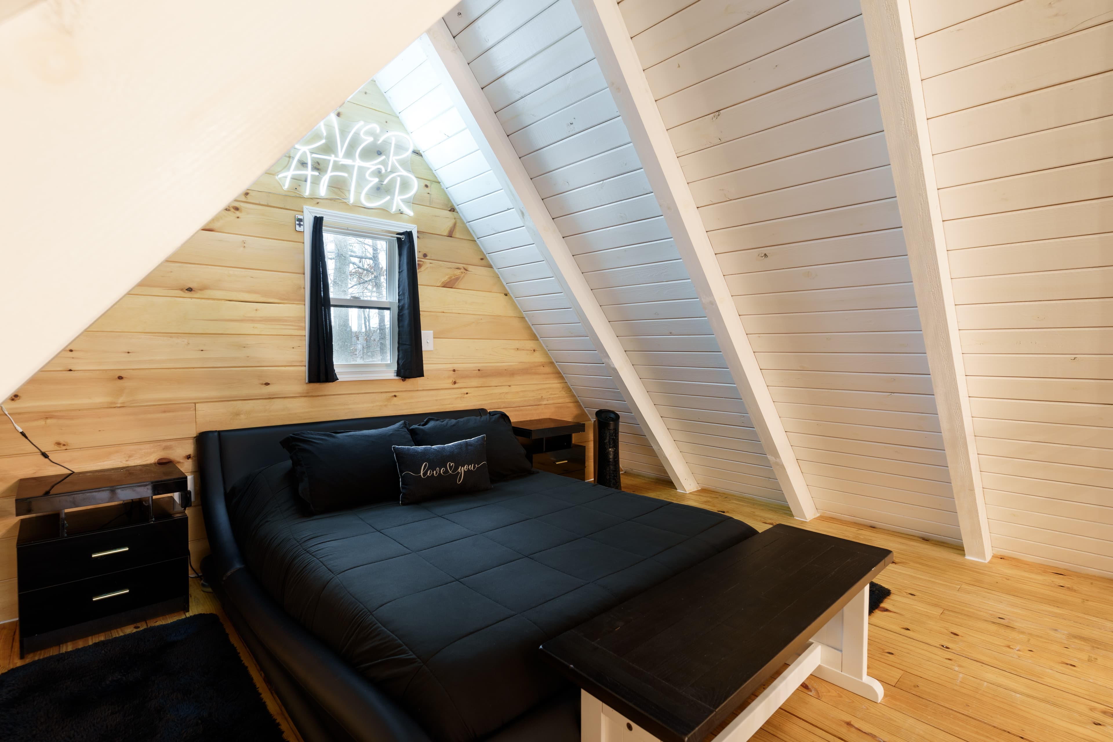 A cozy attic bedroom featuring a black bed, wooden walls, and a neon sign.