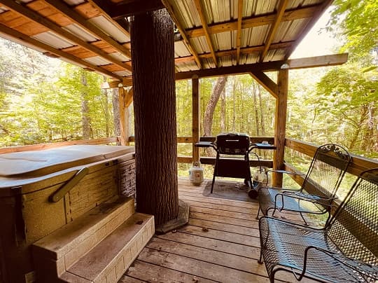 A wooden porch features a hot tub, a grill, and metal chairs surrounded by trees.