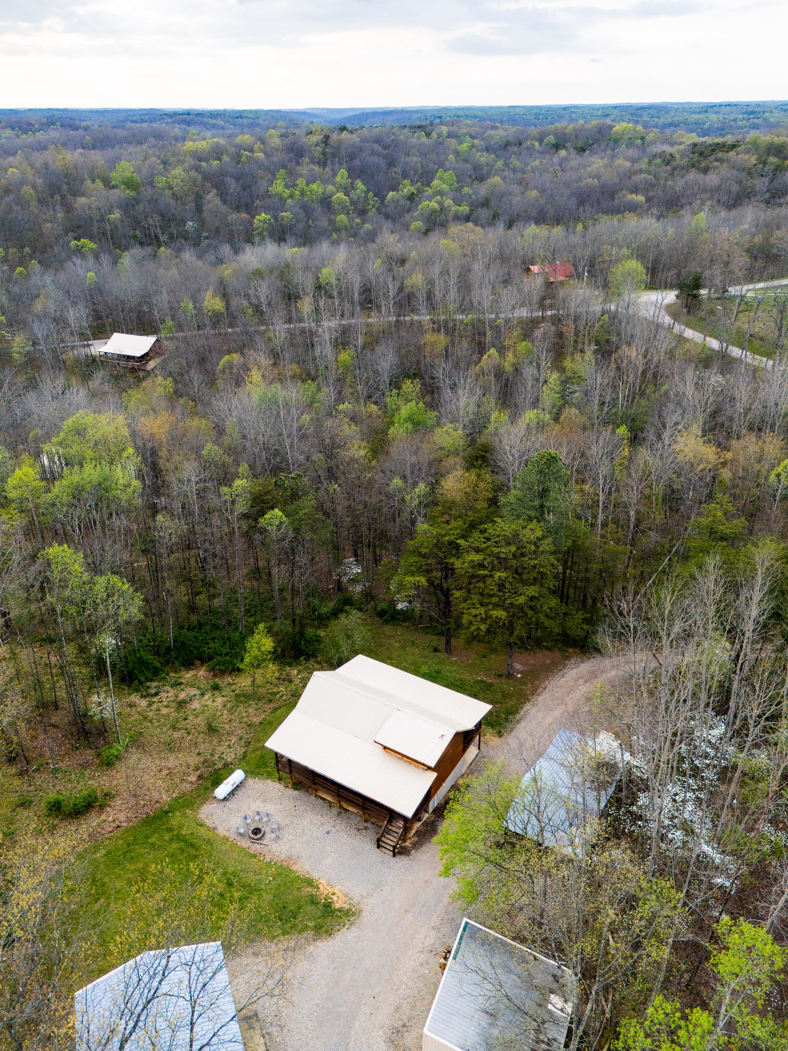 Aerial view of a wooded area with a cabin and several structures along a winding dirt road.