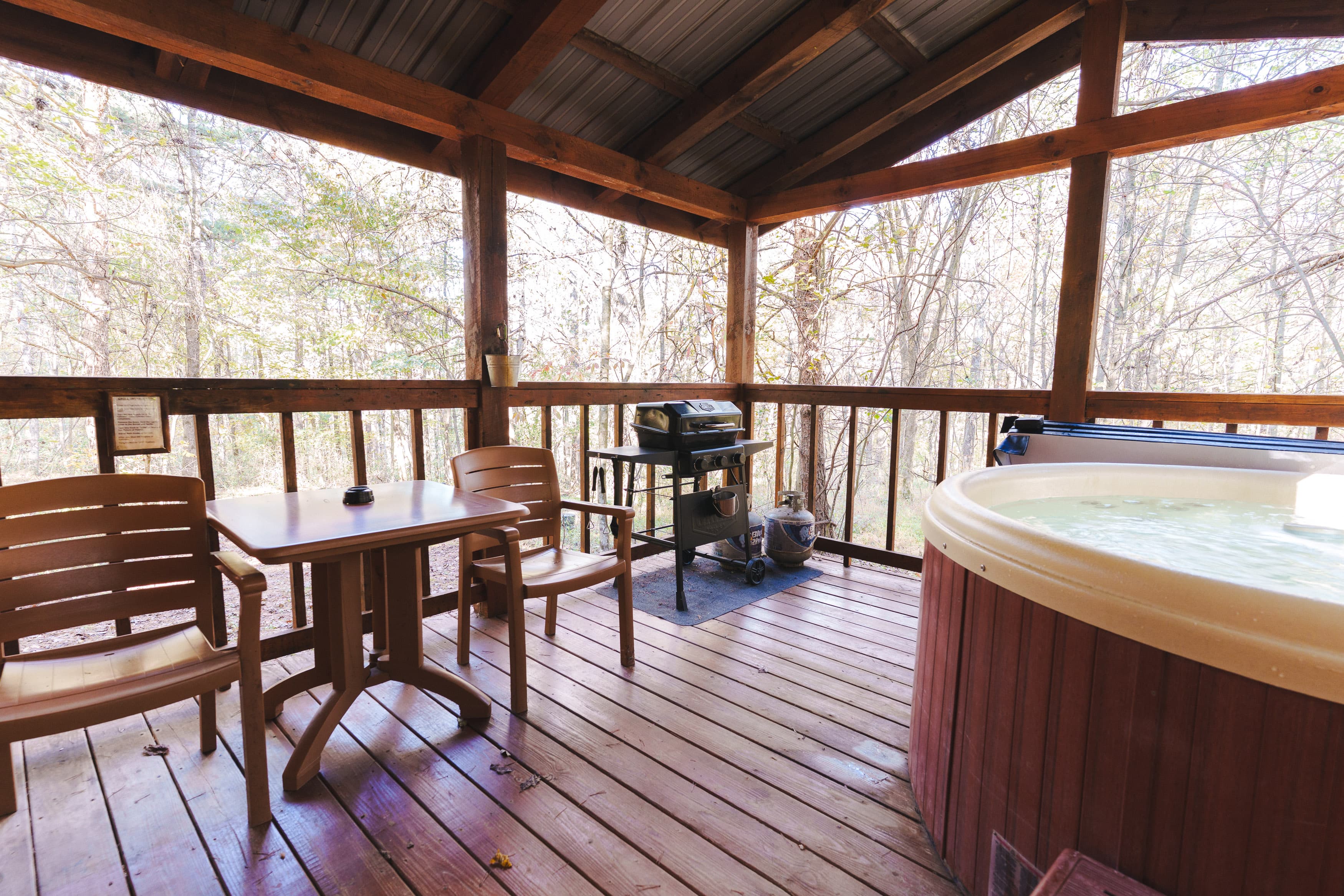 A wooden deck featuring a hot tub, a small table with chairs, and a grill, surrounded by trees.