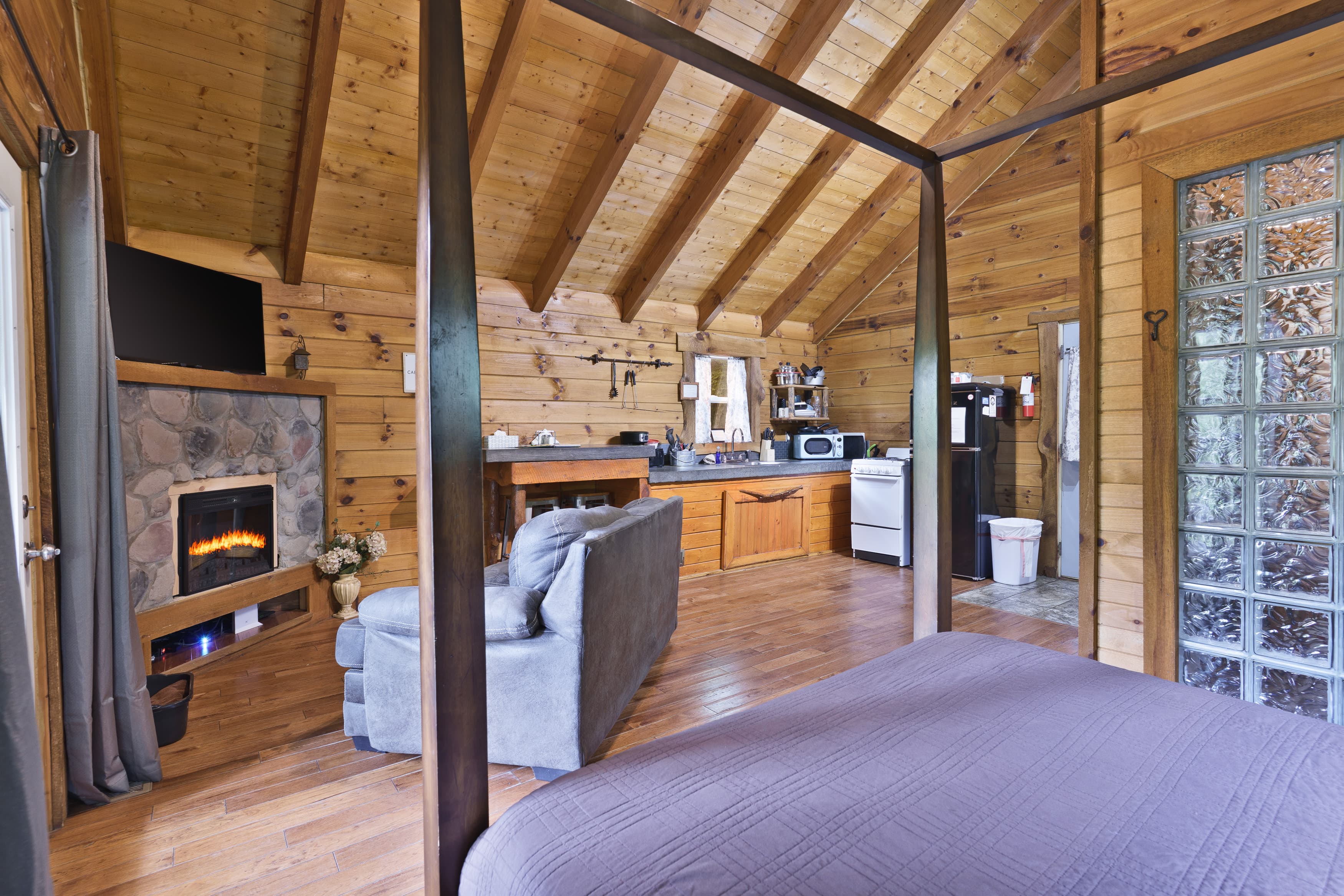 Cozy wooden cabin interior featuring a bed with a view of a living area, kitchenette, and fireplace.