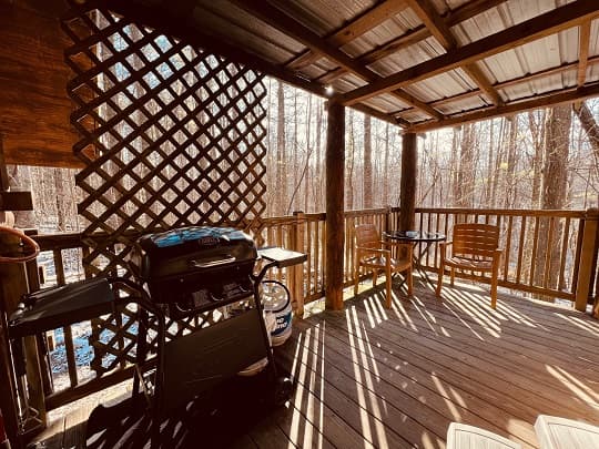 A cozy outdoor deck with a grill, two chairs, and sunlight filtering through trees.
