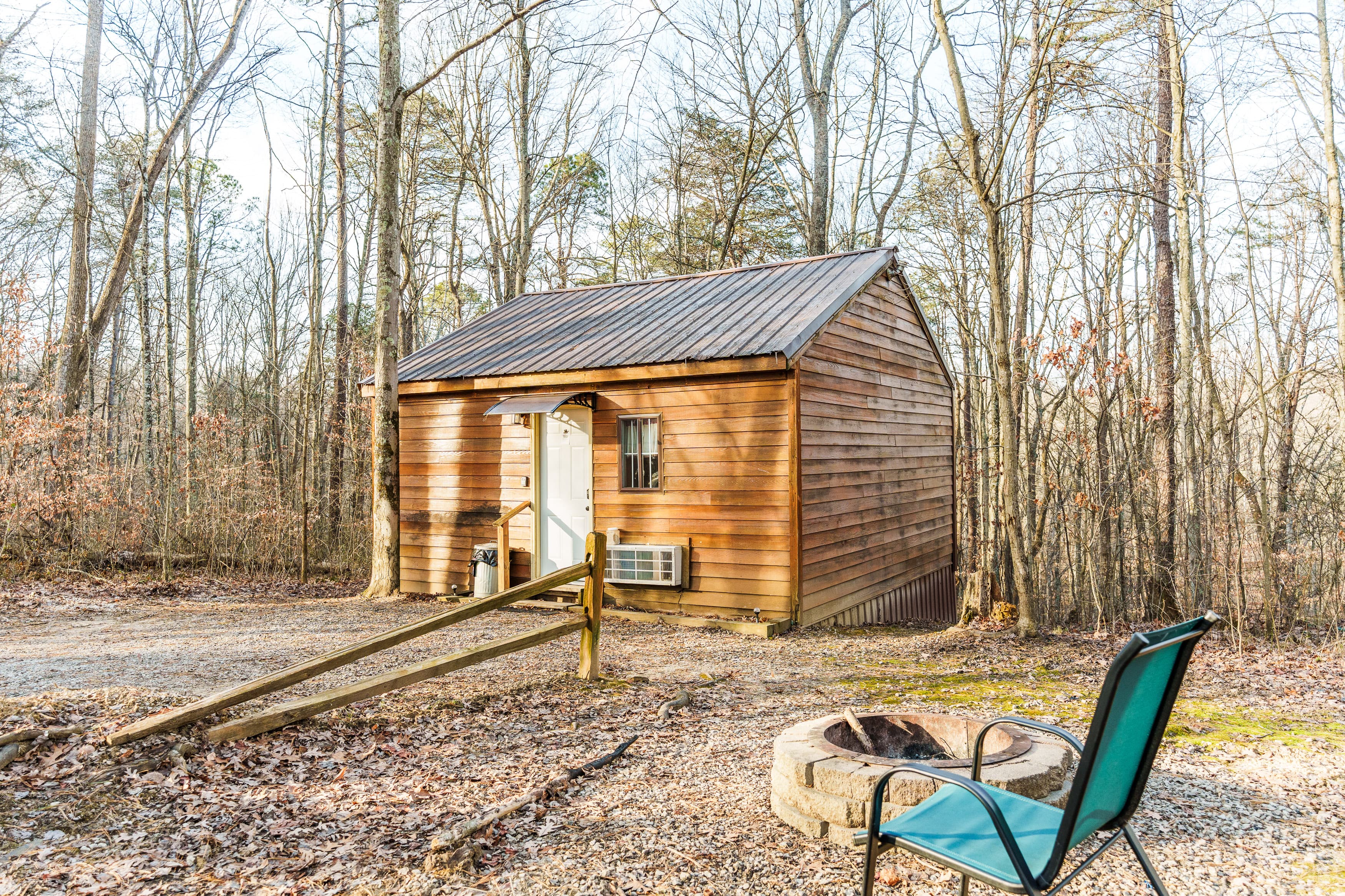 A small wooden cabin surrounded by bare trees, with a fire pit and a chair in front.