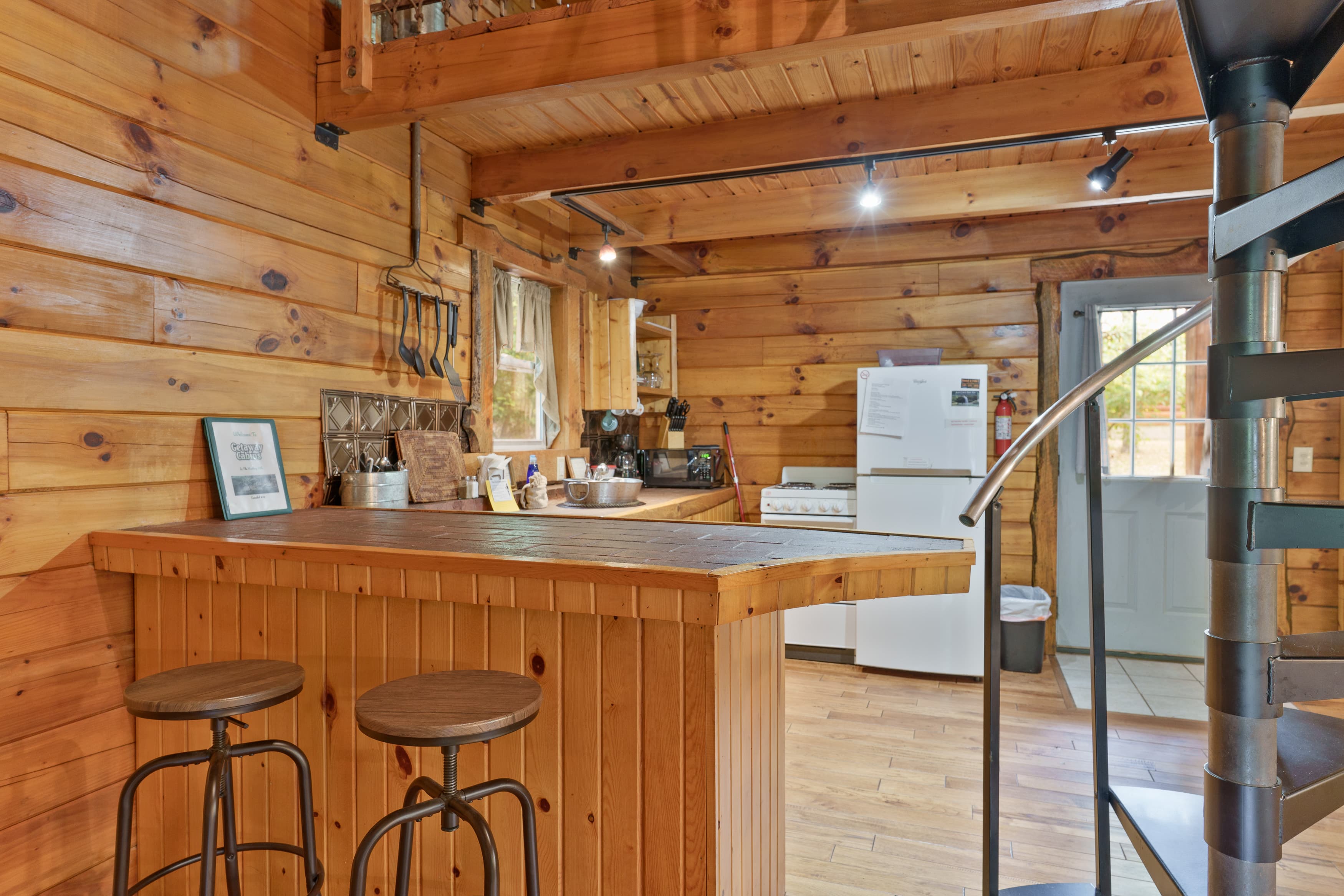 A cozy wooden cabin kitchen with a bar, stools, and a spiral staircase.