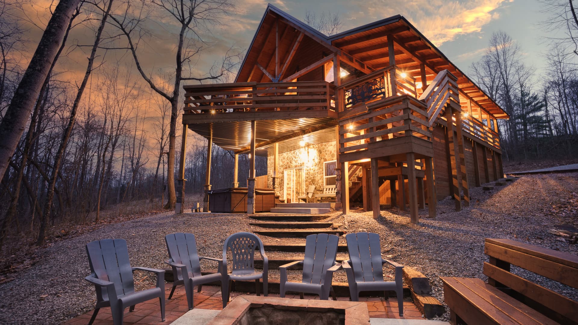 A modern wooden cabin surrounded by trees at dusk, featuring a fire pit and outdoor seating.