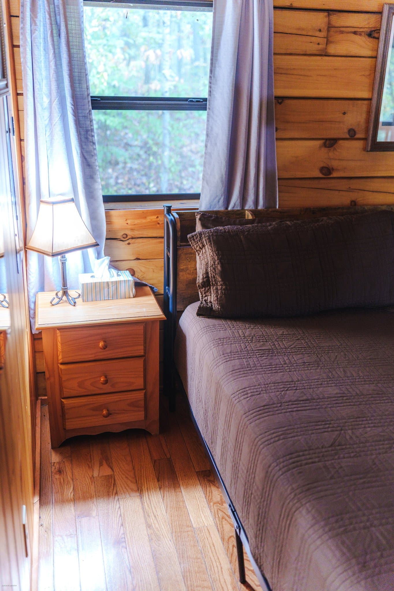 A cozy bedroom featuring a brown quilted bedspread, a wooden bedside table with a lamp and tissues, and large windows with curtains.