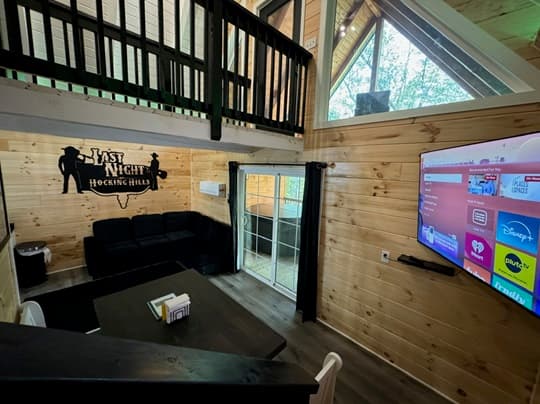 Cozy interior of a wooden cabin featuring a living area with a black couch, dining table, and a mounted TV.