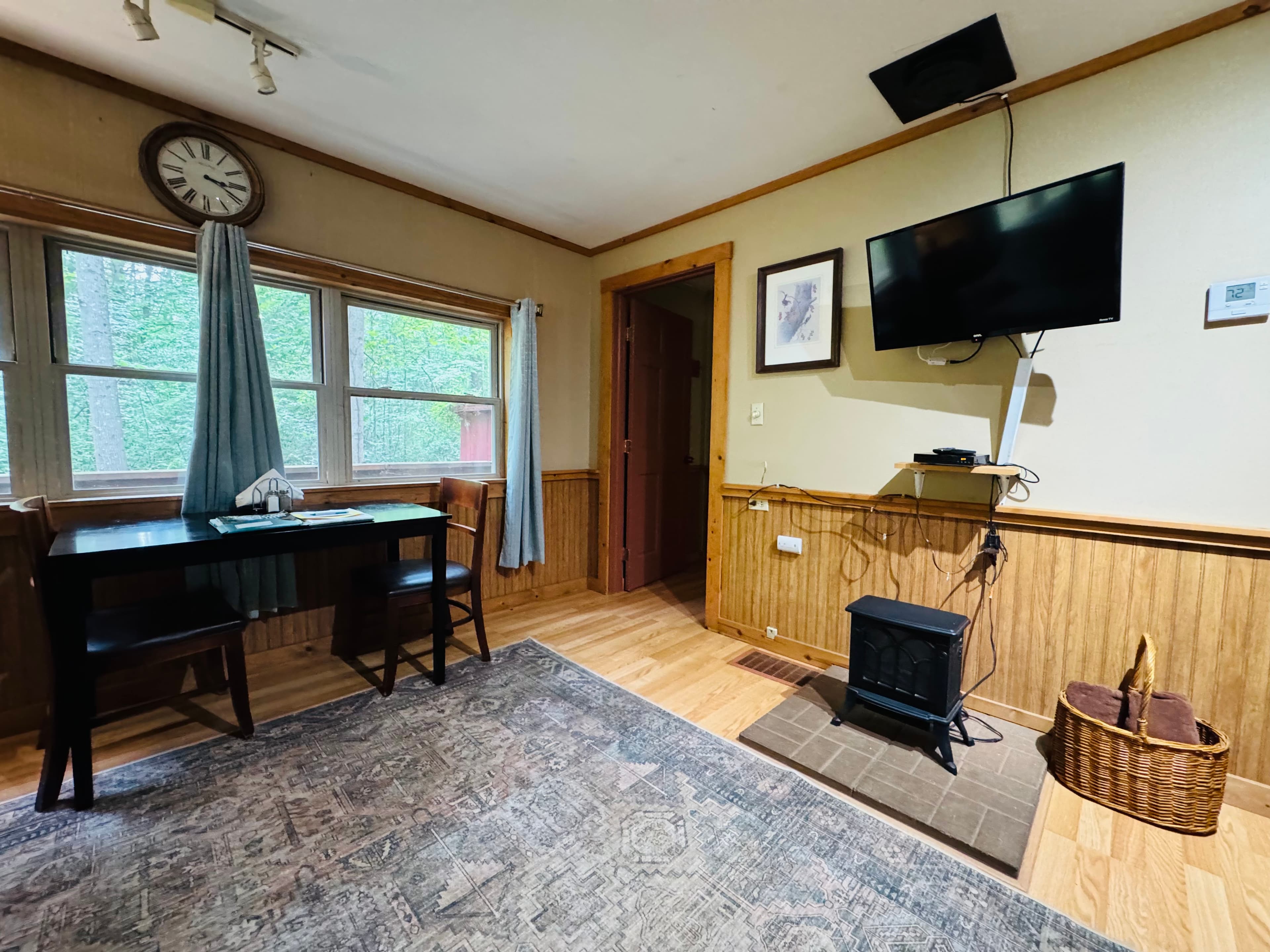 A cozy room featuring a table with chairs, a small stove, a wall-mounted TV, and large windows with curtains.
