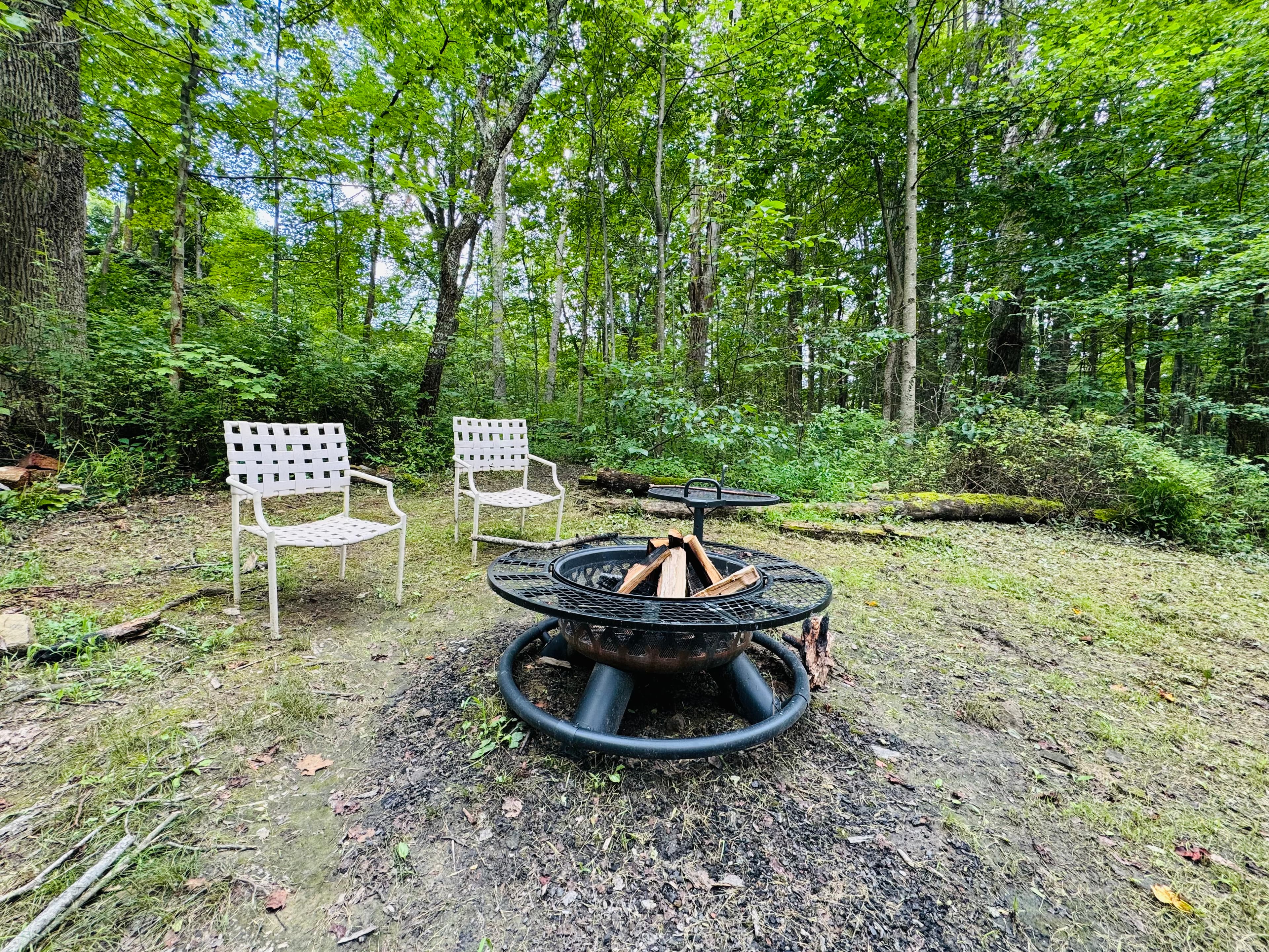 A fire pit surrounded by two white chairs in a wooded area.