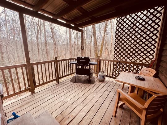 A wooden deck with a grill, chairs, and a view of bare trees in the background.