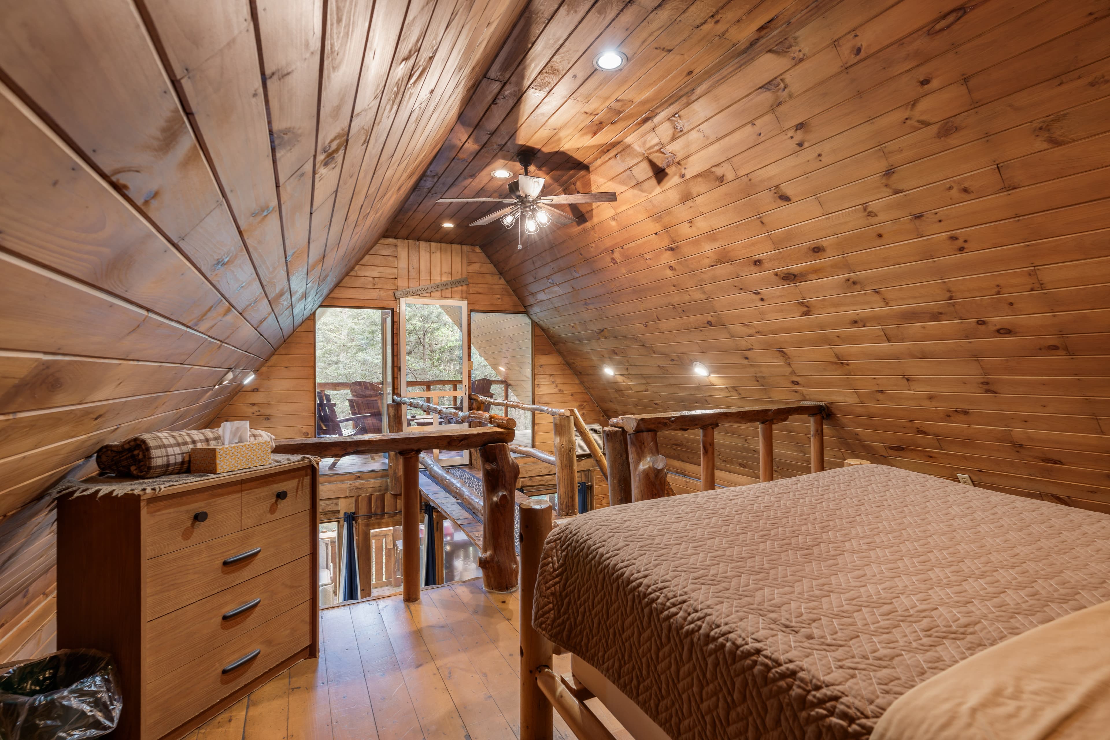 A cozy wooden loft with a bed, dresser, and large windows overlooking nature.
