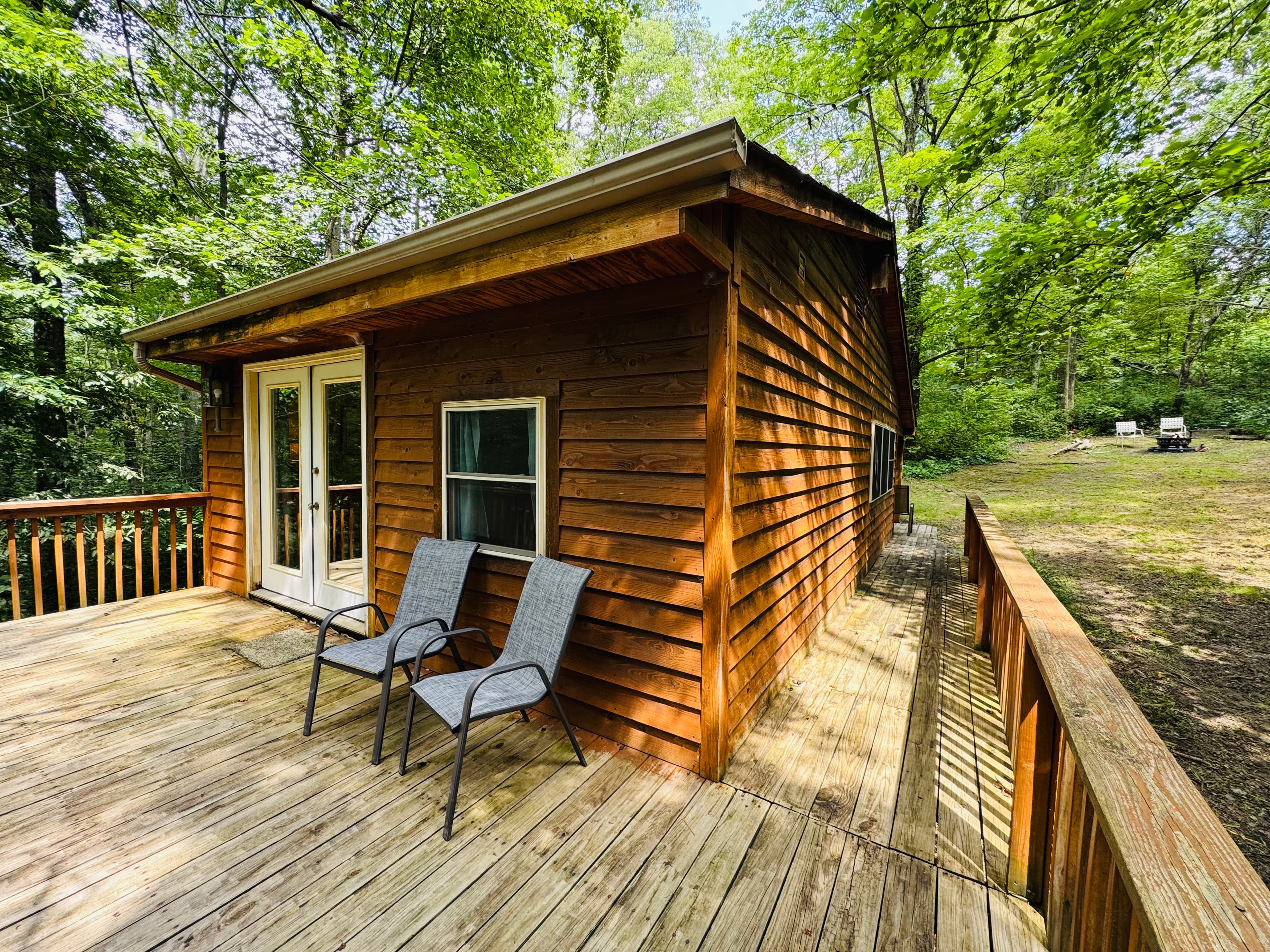 A wooden cabin with a deck and two chairs, surrounded by lush greenery.