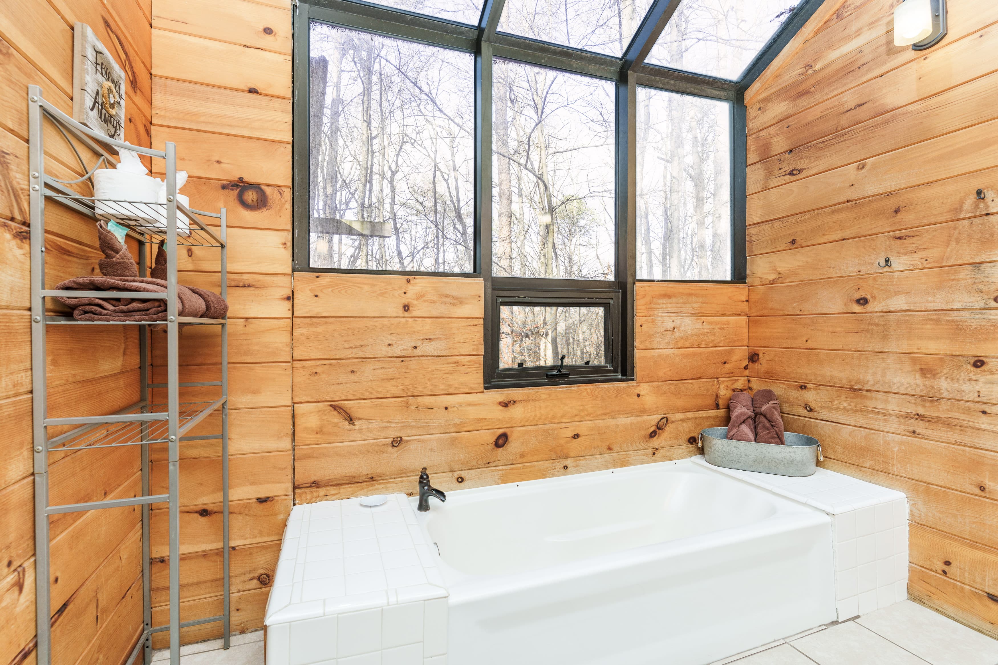 A bright bathroom featuring a white bathtub, wooden walls, and large windows overlooking a wooded area.